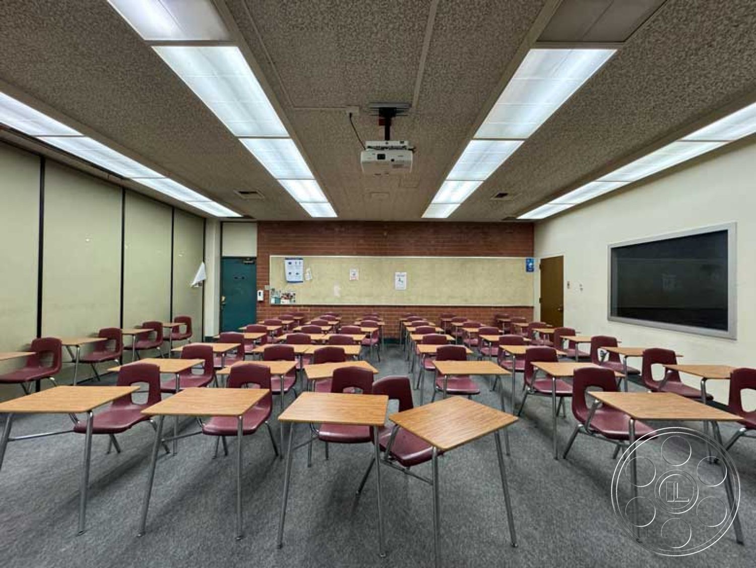 School 3 - carpet flooring, chalkboard wall, exposed brick accent wall, fluorescent ceiling lights, interior classroom space, projector mounted on ceiling, acoustic drop ceiling tiles, metal frame desks, vinyl-covered chairs, paper notice board, dual entrance doors, classroom layout with rows of desks