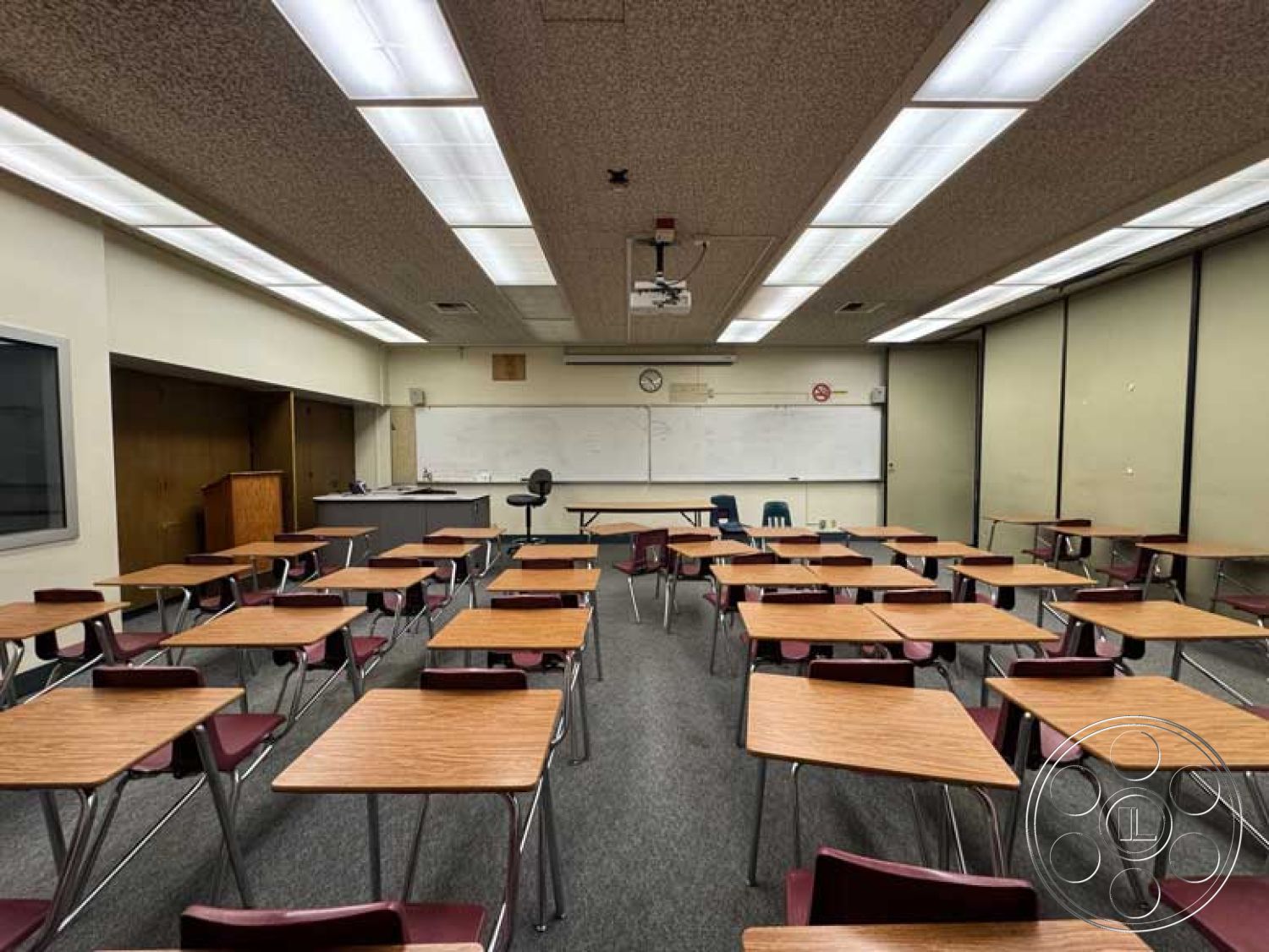 School 3 - exposed ceiling structure, educational environment, acoustic ceiling tiles, fluorescent lighting fixtures, interior classroom, metal chair legs, dry erase board wall treatment, wooden teacher's desk, projector mounted ceiling, classroom layout with rows of desks, carpet flooring classroom, wood laminate desks, blackboard wall treatment, neutral colored walls