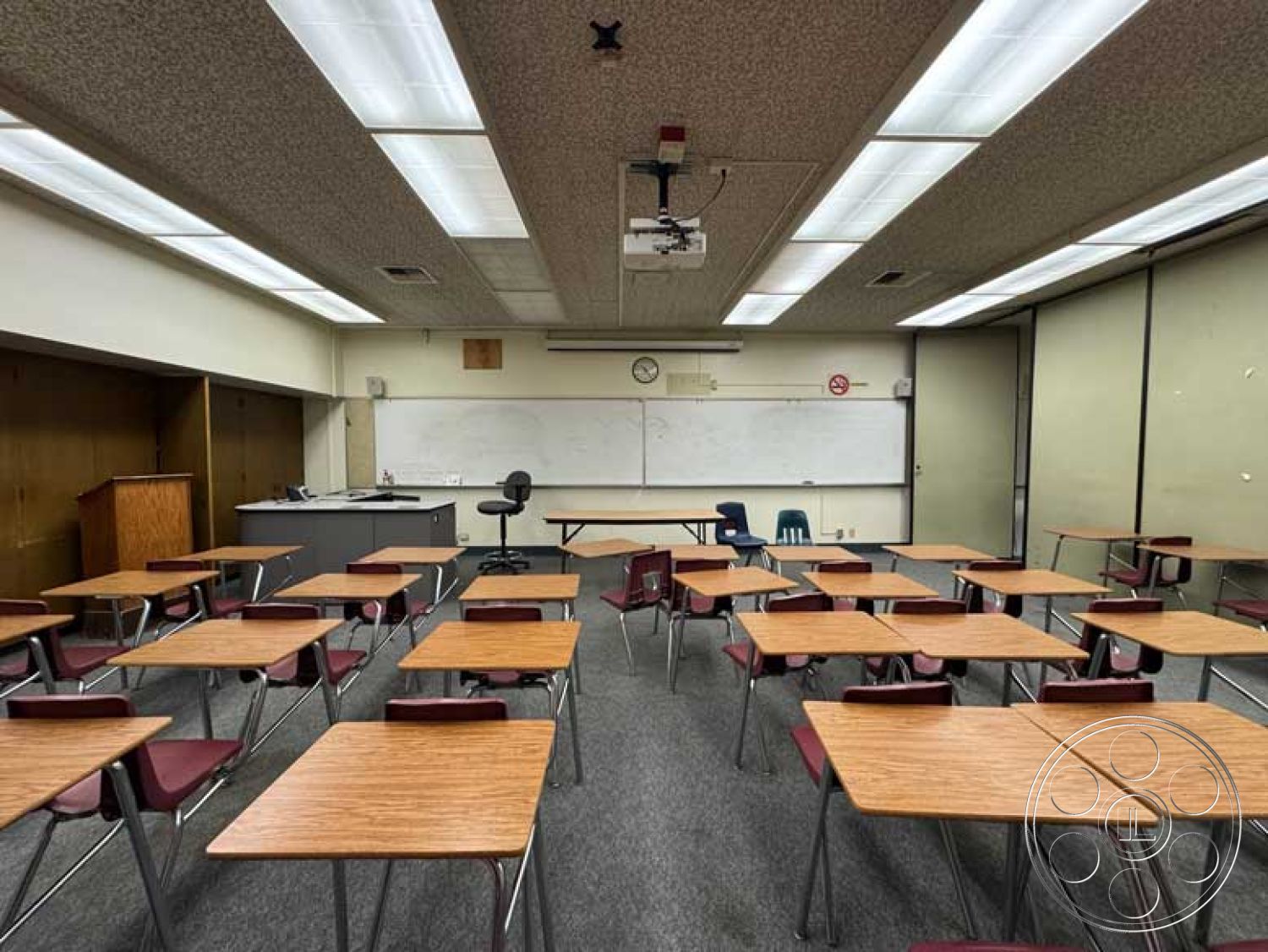 School 3 - open floor plan, carpet flooring, built-in cabinetry, acoustic ceiling tiles, neutral painted walls, fluorescent ceiling lights, whiteboard wall, interior classroom setting, wood laminate desks, metal-framed chairs, corner chalkboard wall, projector mounting, clock mounted on wall, fire safety sign