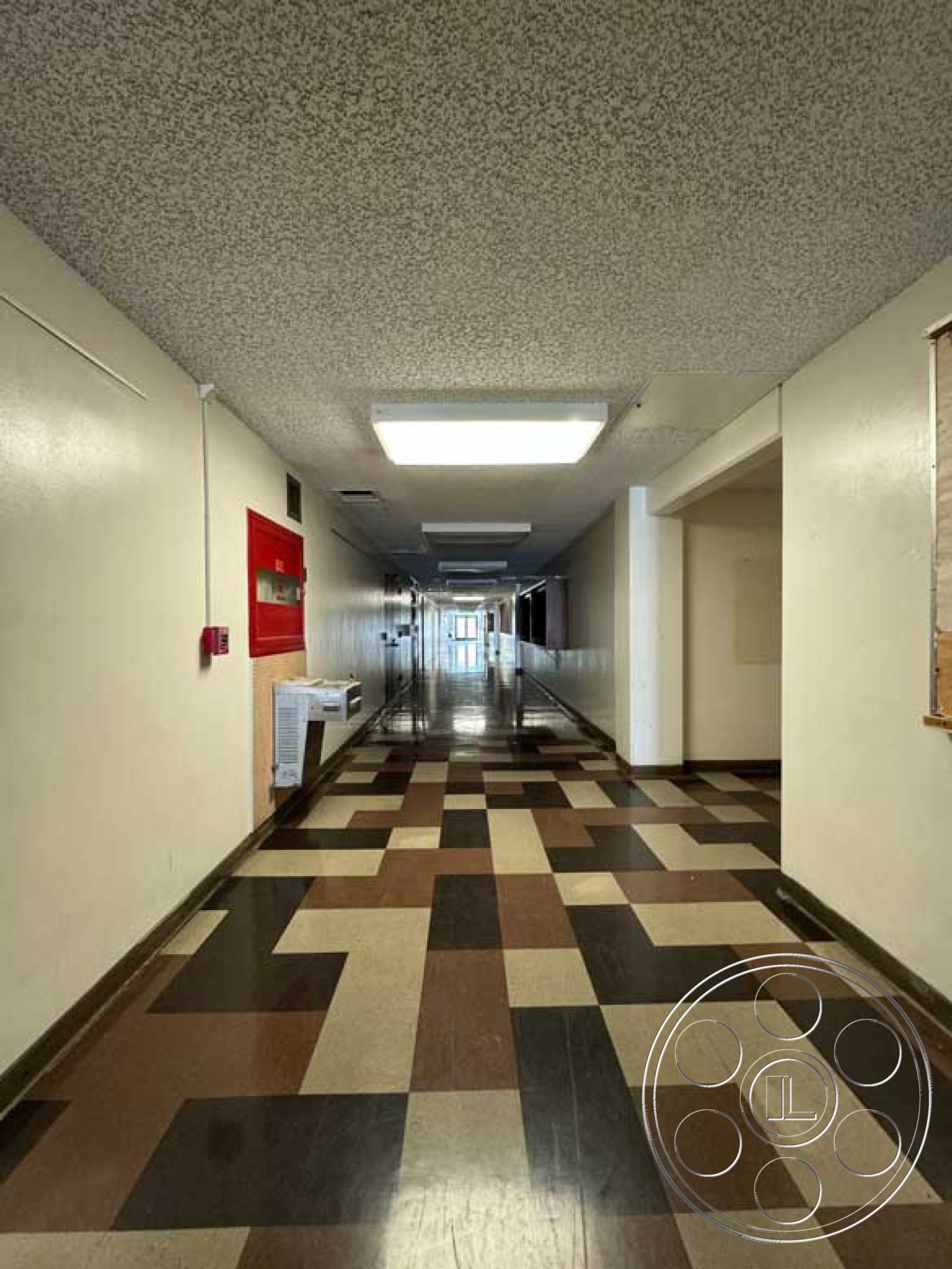 School 3 - tiled flooring, functional design elements, white painted walls, interior corridor, textured ceiling finish, wide corridor design, emergency exit signage, checkerboard tile pattern, flurorescent ceiling lights, brown and cream color tiles, simple wall treatments, fire alarm system mounted on wall, light-colored hallway, exposed structure elements, inward-opening doorways, natural light flooding in from end