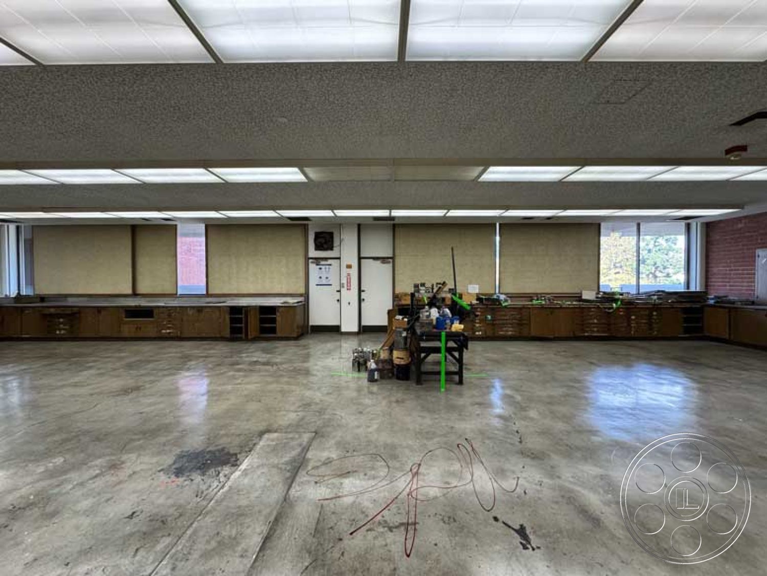 School 3 - industrial lighting fixtures, large floor-to-ceiling windows, exposed concrete floor, minimalist interior design, bright natural light from windows, neutral painted walls, contemporary workspace, open workspace layout, functional workshop environment, flat ceiling with acoustic tiles, wooden cabinetry with drawer storage, large unobstructed interior space, workbench area with tools, urban educational facility interior
