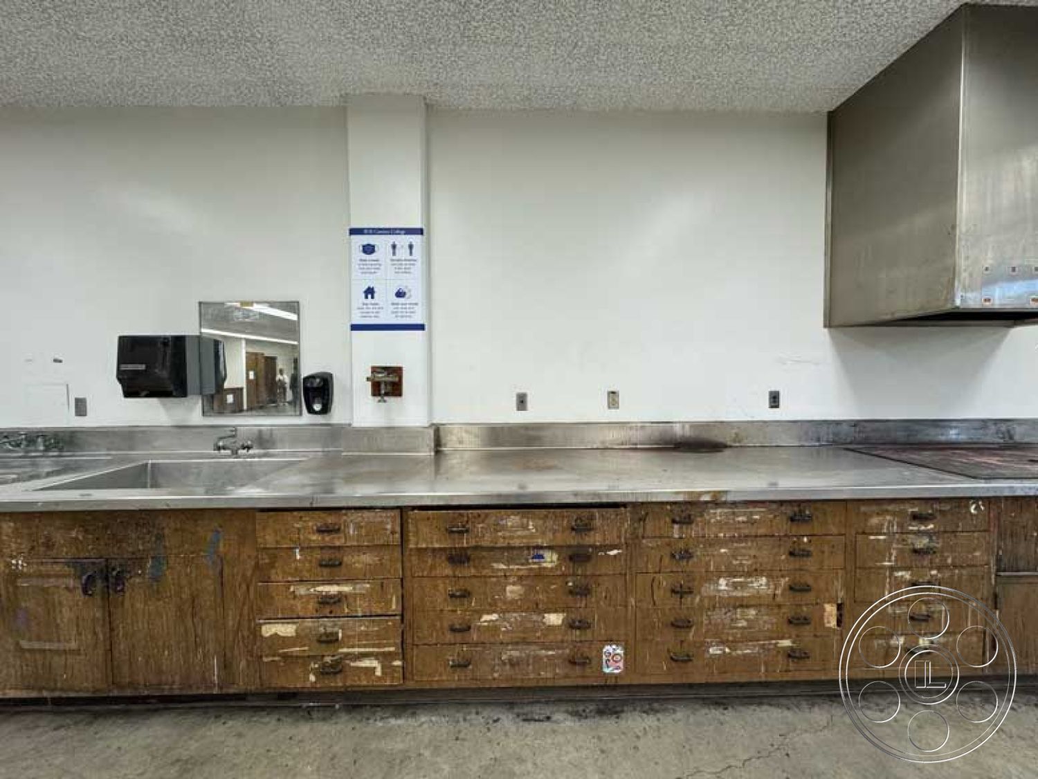 School 3 - interior functional kitchen space, interior polished concrete floor, interior painted drywall walls, interior stainless steel countertops, interior wooden kitchen cabinets, interior industrial-style sink, interior textured acoustic ceiling, interior commercial kitchen layout, interior hardwood drawer fronts, interior stainless steel backsplash, interior utility wall-mounted dispenser, interior illuminated signage, interior open layout workspace, interior practical food preparation area, interior multipurpose kitchen cabinetry