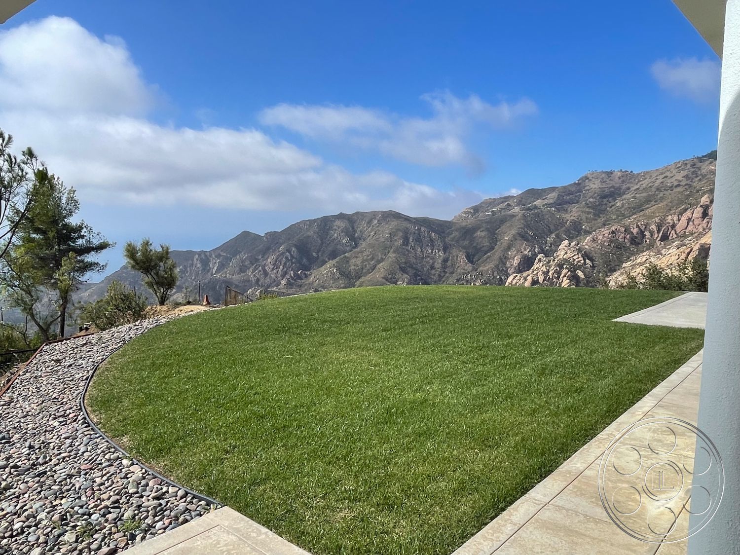 Modern 56 - mountain view, clear blue sky, modern outdoor space, sunny outdoor environment, exterior grass lawn, landscape stones pathway, concrete patio edge, open hillside setting, curved gravel path, natural rock formations, distant horizon vista, tree-lined border