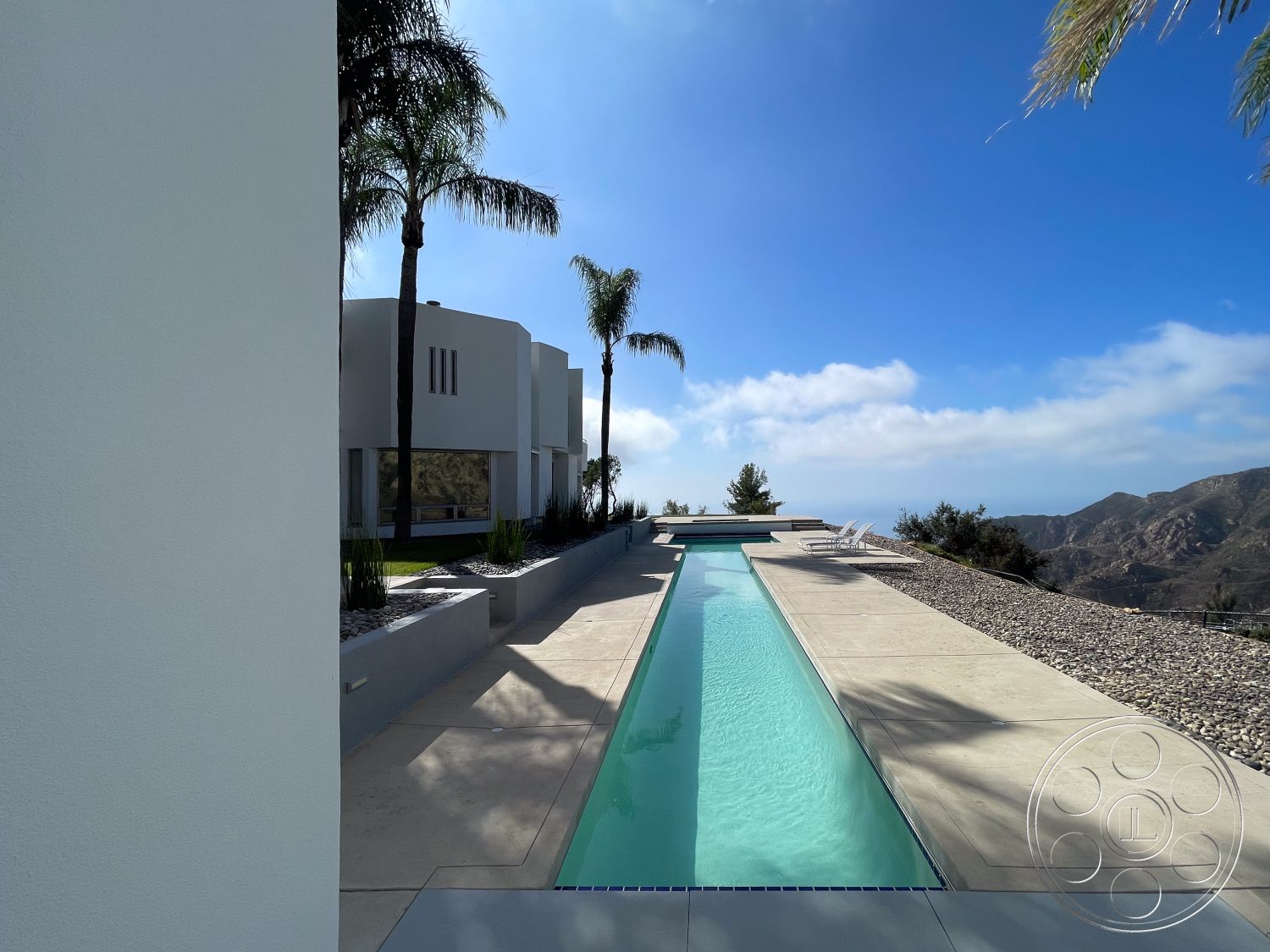 Modern 56 - palm trees, modern landscape design, open outdoor space, mountain view, large floor-to-ceiling windows, luxury outdoor space, concrete pool deck, stucco exterior walls, minimalist architectural style, sunny outdoor seating area, clean lines and geometric shapes, exterior pool area, rectangular infinity pool, light-colored concrete pavers, succulent garden borders, stone gravel landscaping, skyline backdrop