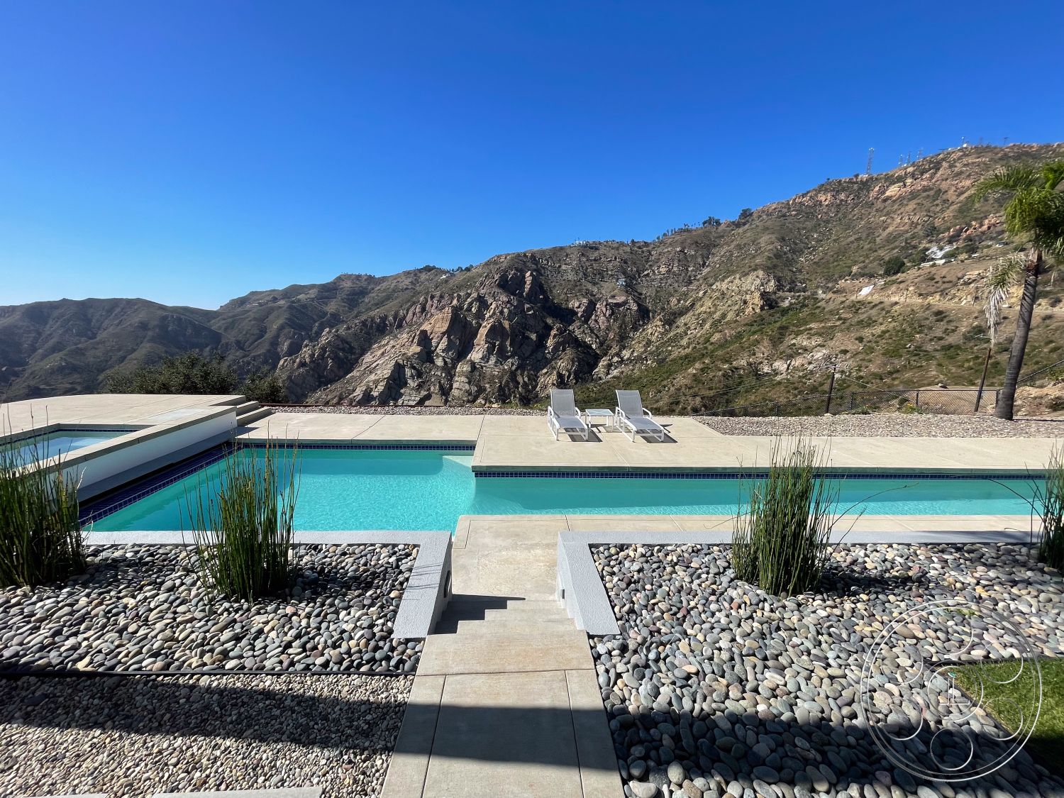 Modern 56 - clear blue sky, outdoor leisure area, drought-tolerant landscaping, minimalist outdoor space, concrete patio flooring, concrete steps leading to pool, exterior concrete pool deck, rectangular infinity pool, glass pool edge, modern outdoor architecture, landscaped rock garden, surrounding mountain view, low-maintenance pebble stone, sun loungers by pool, modern pool design, expansive mountain backdrop