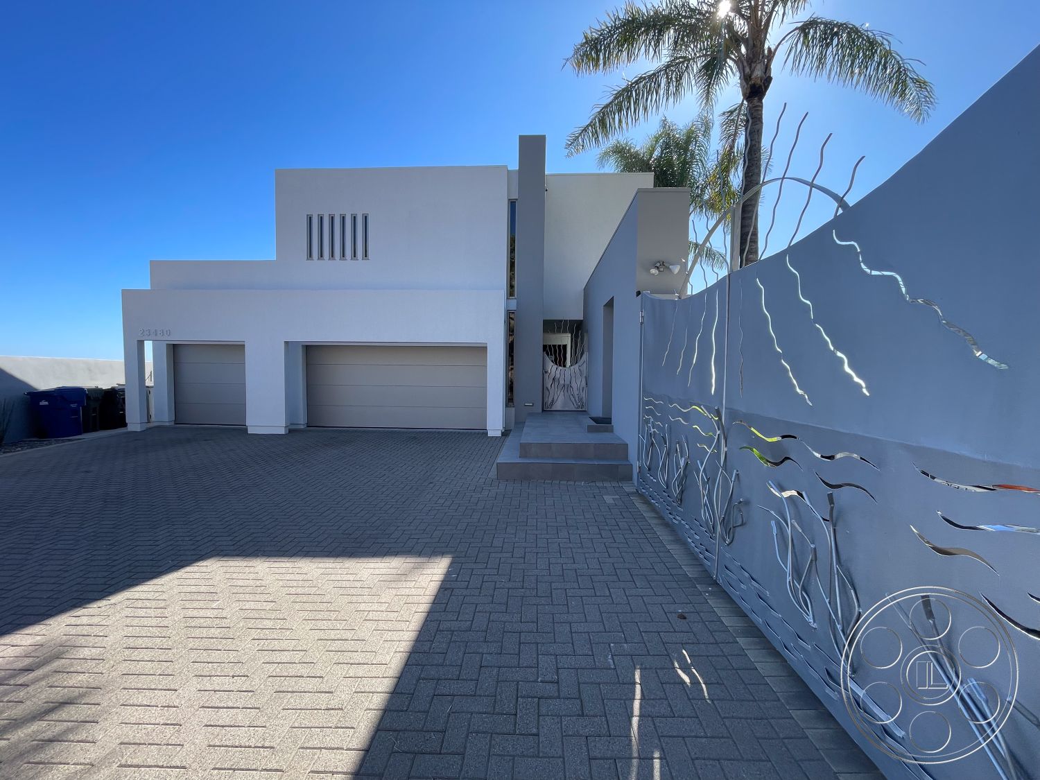 Modern 56 - large windows, exterior, paved driveway, modern landscaping, symmetrical facade, open parking area, flat roof design, stucco exterior walls, palm tree landscaping, minimalist facade, textured plaster wall details, glass entry doors, smooth concrete steps, geometric architectural shapes, interlocking brick driveway, decorative metal gate, white painted garage doors, horizon view, light gray exterior finish