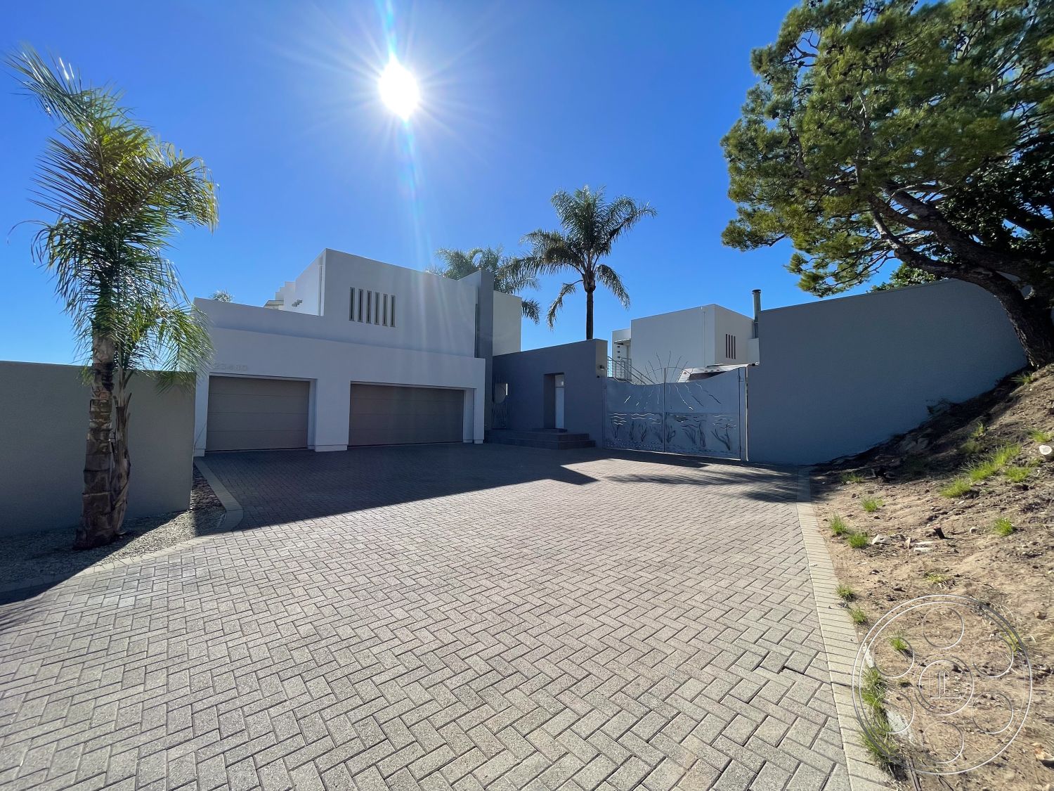 Modern 56 - stucco wall finish, clean lines and geometric shapes, landscaped with palm trees, exterior lighting fixtures, paved driveway with interlocking brick pavers, modern two-story home exterior, contemporary concrete entrance gate, desert environment setting, materials: concrete and brick, expansive blue sky backdrop, exterior of house with angular rooflines, stylish garage door design, modern minimalist architectural style, elevated site with hillside landscaping