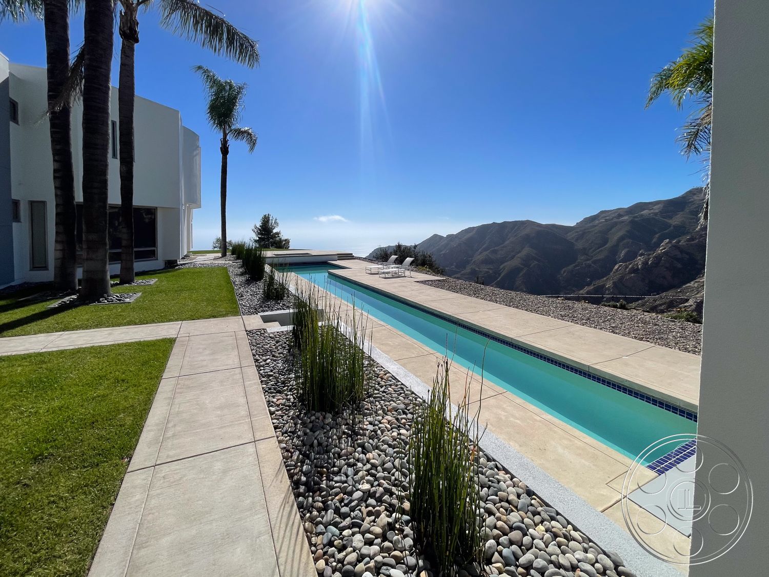 Modern 56 - exterior, large glass windows, sloped terrain, infinity pool, rectangular pool, mountain view setting, concrete patio flooring, contemporary home design, concrete pool border, geometric shapes in architecture, open sky view, spacious outdoor living space, palm tree accents, sun-drenched terrace, minimalist outdoor seating area, smooth white stucco walls, textured stone deck, landscape rock garden, native grass landscaping, serene natural environment