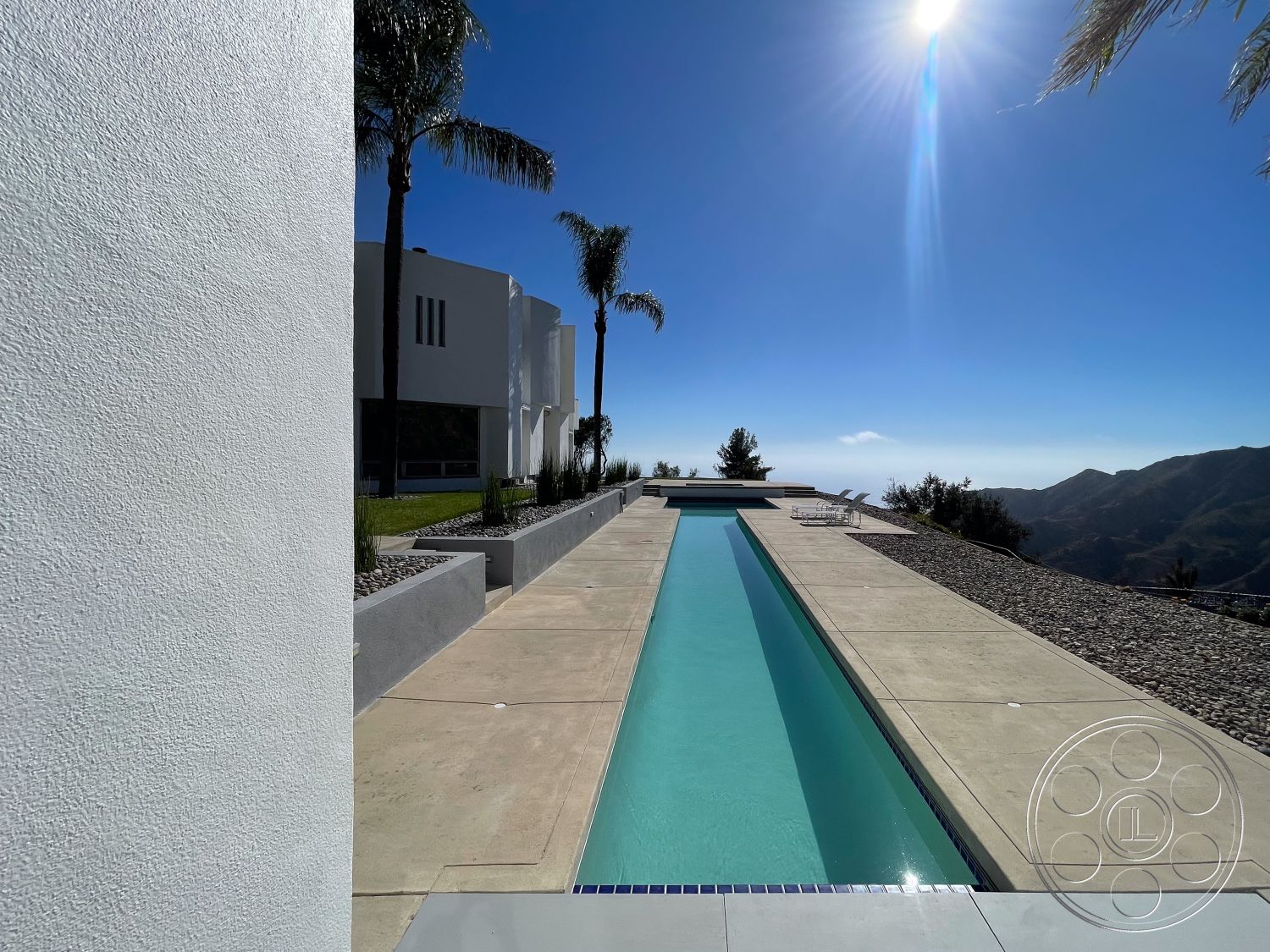 Modern 56 - exterior, white stucco walls, outdoor seating area, floor-to-ceiling windows, rectangular pool, minimalist architecture, sunny exterior, mountain view setting, landscaped garden area, concrete patio flooring, concrete pool border, palm tree accents, modern outdoor living space, smooth plaster wall, ADA-compliant pool access, landscape gravel pathway, luxury residential property, swimming pool with water feature, sleek poolside design