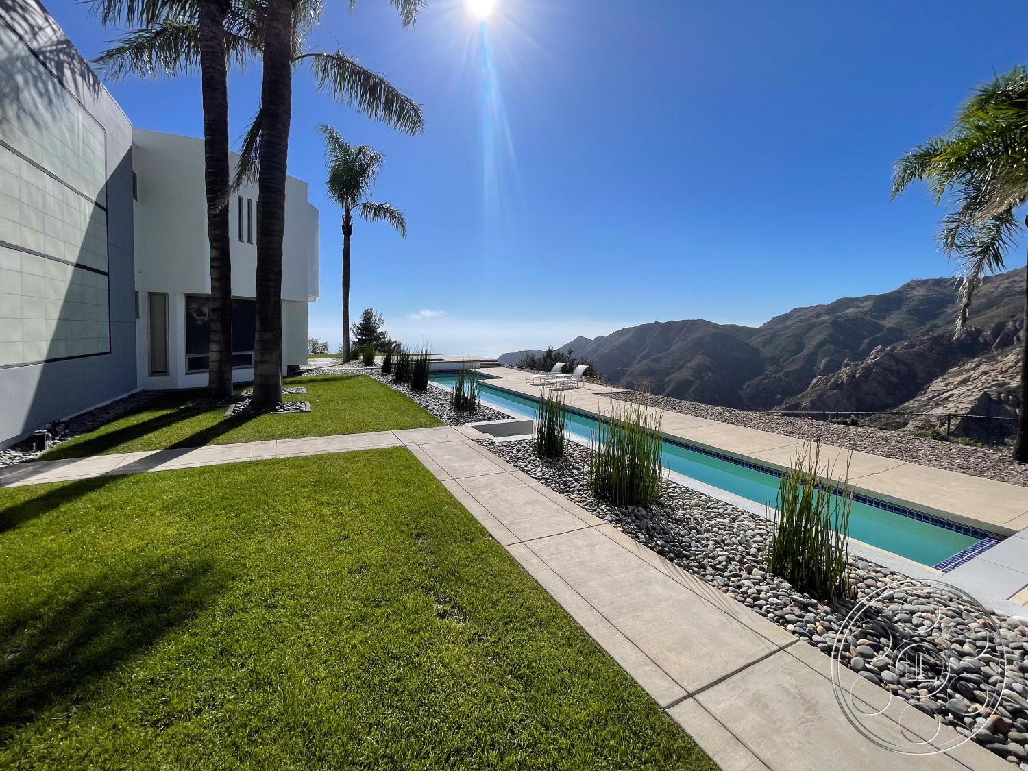 Modern 56 - open space design, outdoor living space, lush green lawn, tropical palm trees, rectangular pool, minimalist architecture, large sliding glass doors, stone pool deck, sunny outdoor setting, concrete patio floor, mountain view background, gravel landscape border, modern exterior wall, integrated pool landscaping, planted aquatic grass, exposed concrete walkway