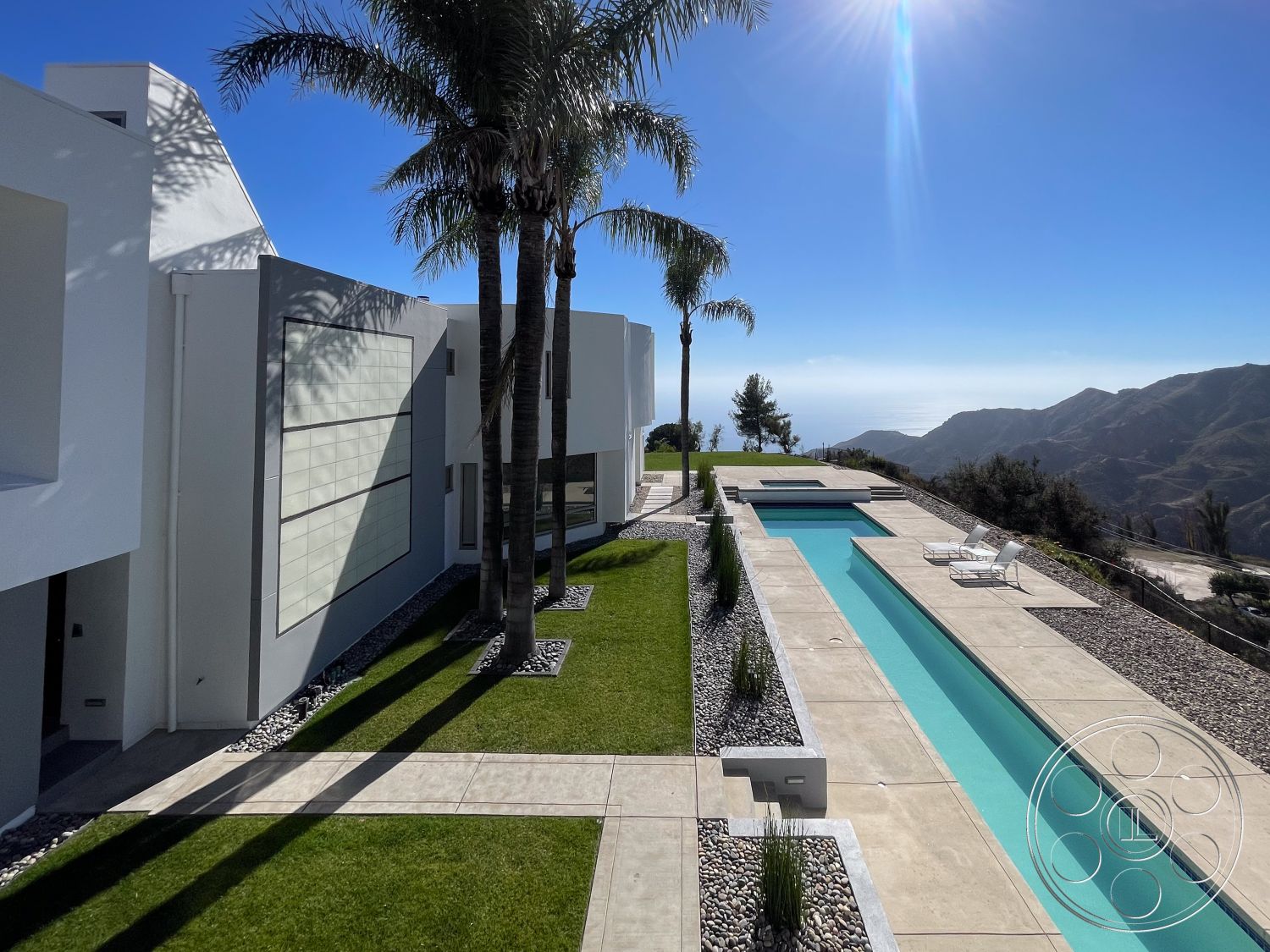 Modern 56 - exterior, white stucco walls, palm trees, clear sky, concrete pathway, outdoor lounge area, landscaped lawn, large floor-to-ceiling windows, modern exterior lighting, flat roof design, mountain view setting, minimalist architectural style, concrete pool coping, rectangular infinity pool, tile pool deck, poolside chaise lounges, stone gravel landscaping, geometric landscaping features, glass patio railing