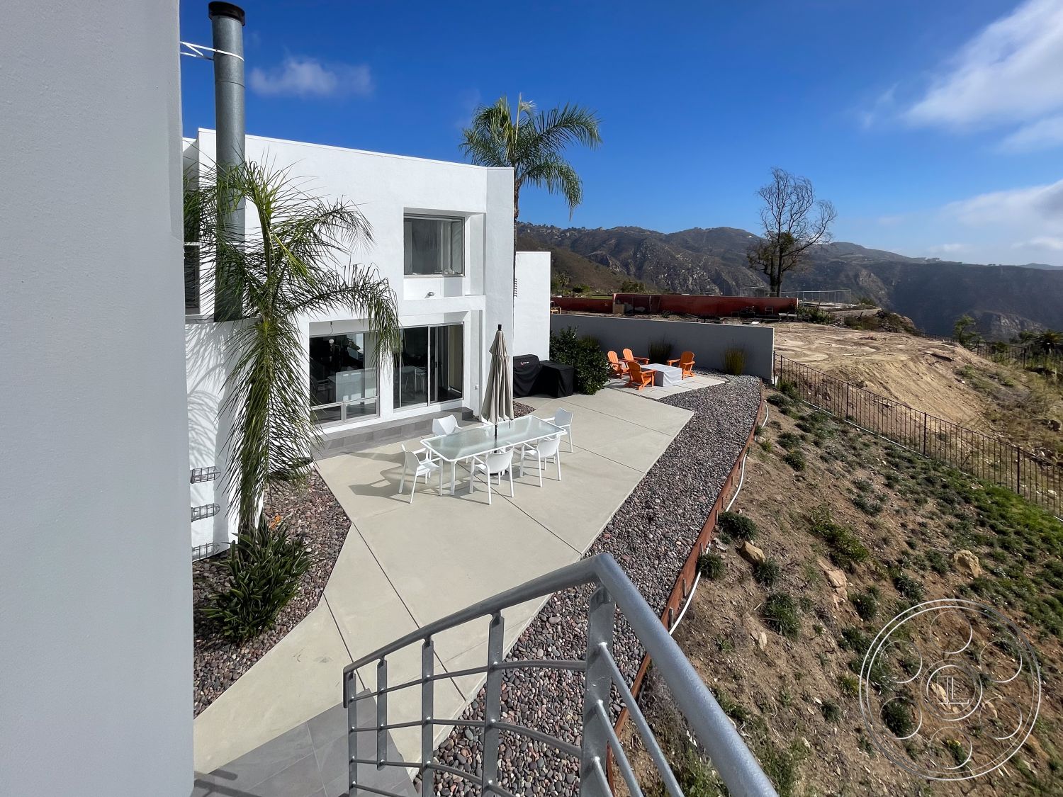 Modern 56 - exterior, white stucco walls, modern outdoor furniture, smooth concrete surface, floor-to-ceiling windows, sunny outdoor area, mountain view setting, minimalist architectural style, concrete patio floor, palm trees landscaping, rectangular patio, landscape gravel border, cylindrical metal chimney, open hillside location, hardscape with vegetation