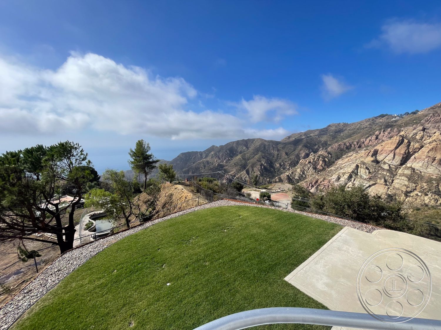 Modern 56 - exterior, landscaped garden, sloped terrain, stone retaining wall, concrete patio flooring, grassy lawn area, natural hillside backdrop, clear blue skies, mountainous landscape view, circular gravel pathway, freestanding trees, residential property setting