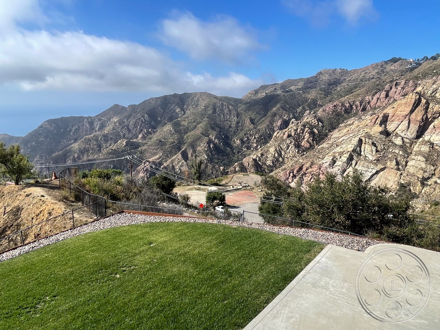 Modern 56 - exterior, lush greenery, curved driveway, outdoor living area, sunny outdoor space, hilly terrain, scenic vista, urban edge, mountain view setting, concrete patio floor, open sky view, adjacent roadway, natural stone landscaping, gravel border, landscaped lawn area, fenced hillside property, frontage with fencing, drainage gravel base