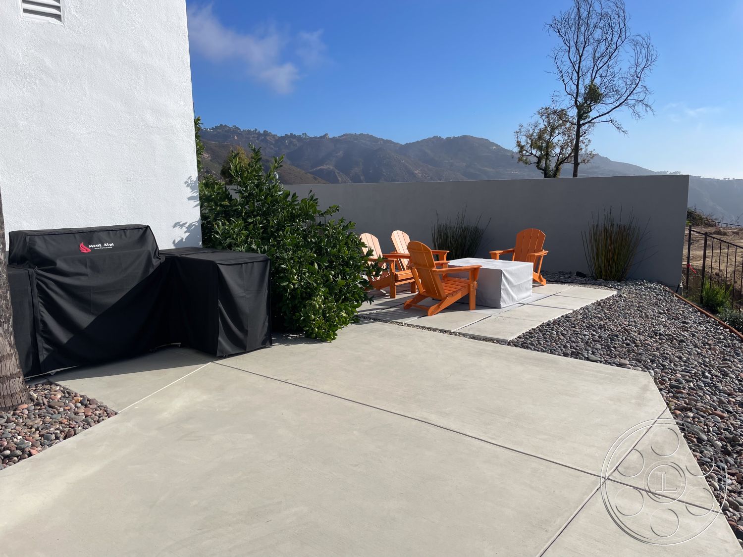 Modern 56 - exterior gravel landscaping, exterior hillside location, exterior mountain view, exterior concrete patio flooring, exterior rectangular concrete patio slab, exterior minimalist white stucco walls, exterior black barbecue grill cover, exterior orange Adirondack chairs, exterior concrete table surface, interior seamless transition from patio to landscape, exterior contemporary outdoor seating area, exterior low maintenance shrubbery, exterior modern fence, exterior natural stone border