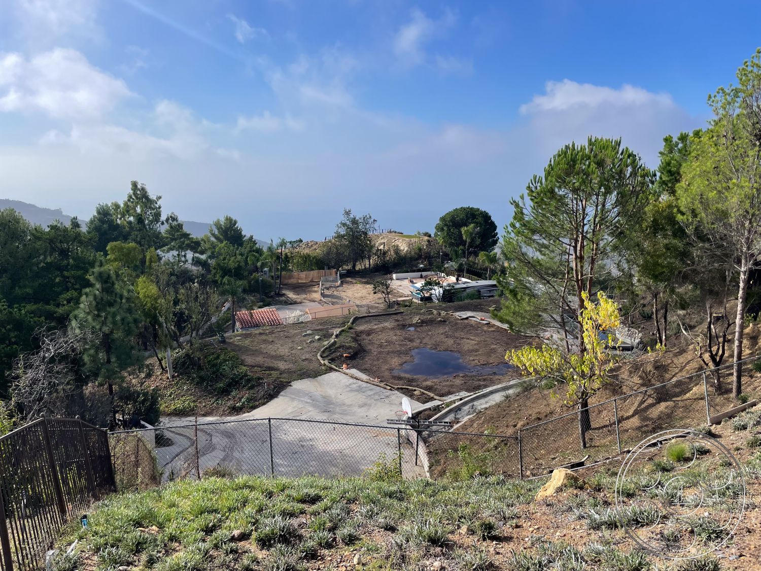 Modern 56 - exterior landscape with unobstructed ocean view, concrete foundation base for future construction, gravel driveway leading to planned home entry, fenced yard with chain-link perimeter fencing, paved path with potential for stone walkway, natural hillside terrain with lush vegetation, open area for garden or pool installation, sloped land for tiered landscaping design, surrounding pine trees providing privacy, future plans for outdoor entertaining space, clear skies indicating coastal climate, future building site in a tranquil setting