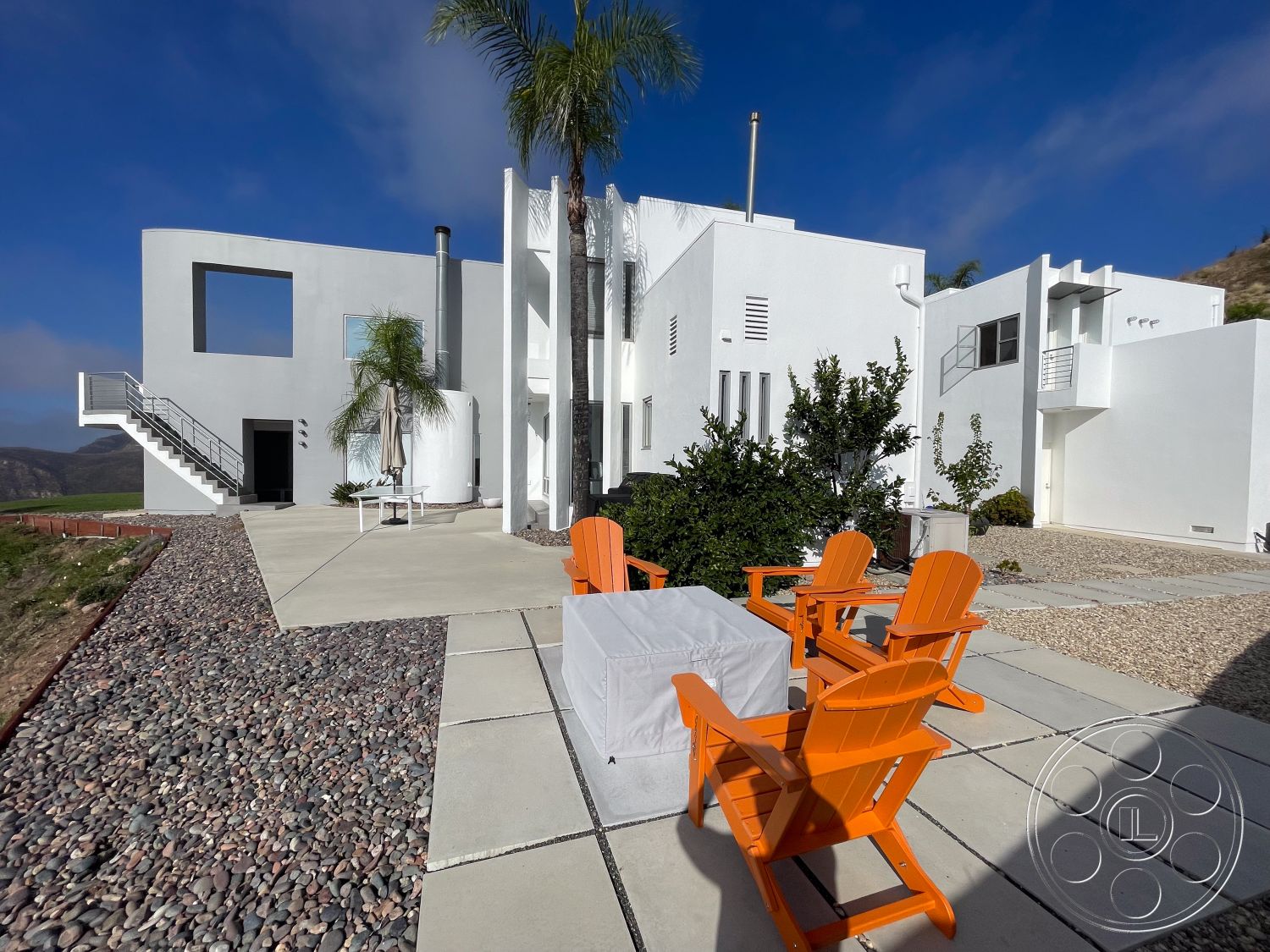 Modern 56 - open outdoor space, contemporary outdoor seating area, green shrub landscaping, pebble stone landscaping, desert environment setting, modern minimalist architectural style, exterior house with white stucco walls, rectangular concrete patio area, stainless steel stair railing, floor-to-ceiling window openings, sloping hillside location, flat concrete outdoor surface, bright orange Adirondack chairs, large geometric window cutout, sunny weather conditions