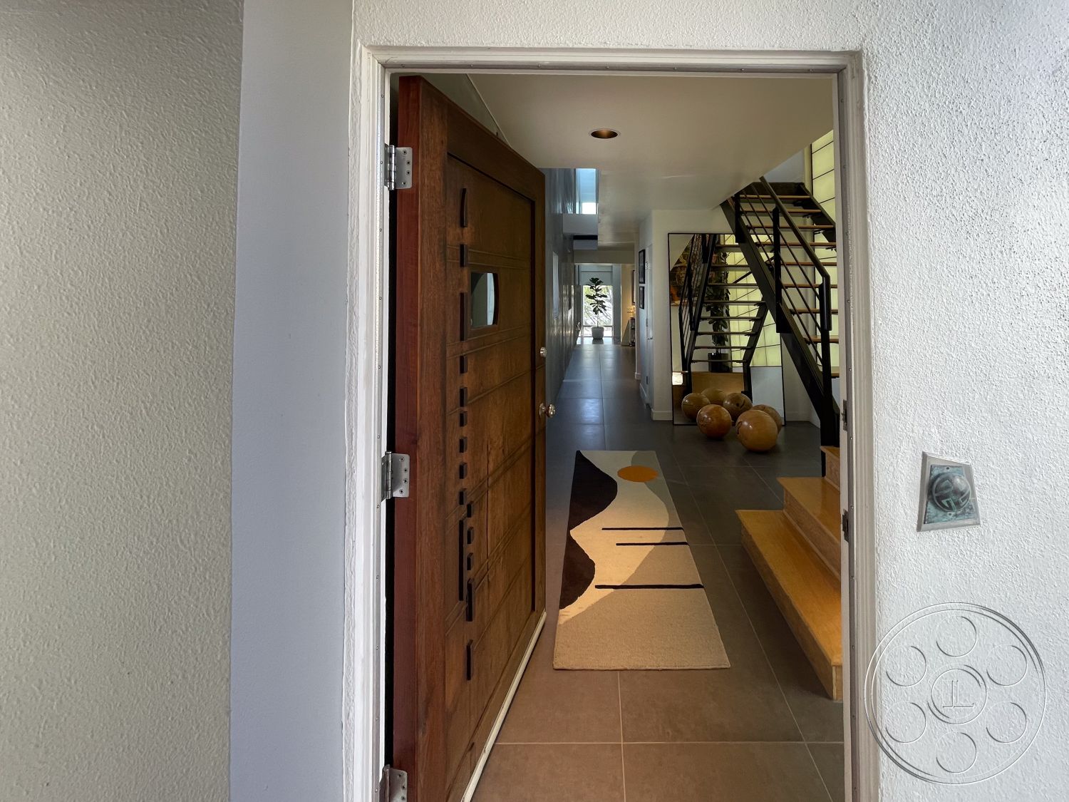 Modern 56 - open floor plan, bright ambiance, natural light from large windows, sculptural lighting fixtures, modern art display, minimalist interior design, textured plaster wall interior, high ceiling entryway, stainless steel stair railings, wood front door with geometric cutouts, gray tile flooring hallway, floating wood steps, contemporary rug with abstract design, exposed stair tread, potted plants accentuating space, internal hallway leading to living area