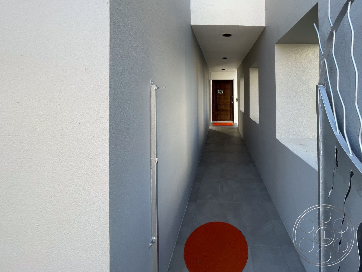 Modern 56 - minimalist design, modern architectural style, wooden front door, recessed ceiling lights, smooth plaster walls, gray ceramic tile flooring, exterior corridor, wide corridor, decorative metal gate, orange circular doormat, window openings