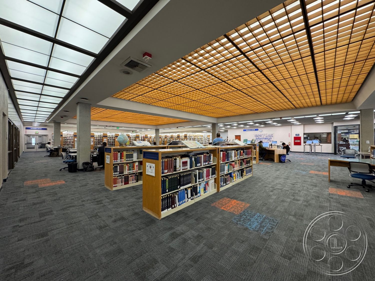School 3 - neutral wall colors, textured carpet flooring, large glass windows for natural light, interior library space, wooden bookshelf units, drop ceiling with grid lighting, ambient glass block ceiling design, open layout reading area, modern study space, informational desk area, multi-functional library design, shared workspace environment, flexible seating arrangements, public resource center, full access layout