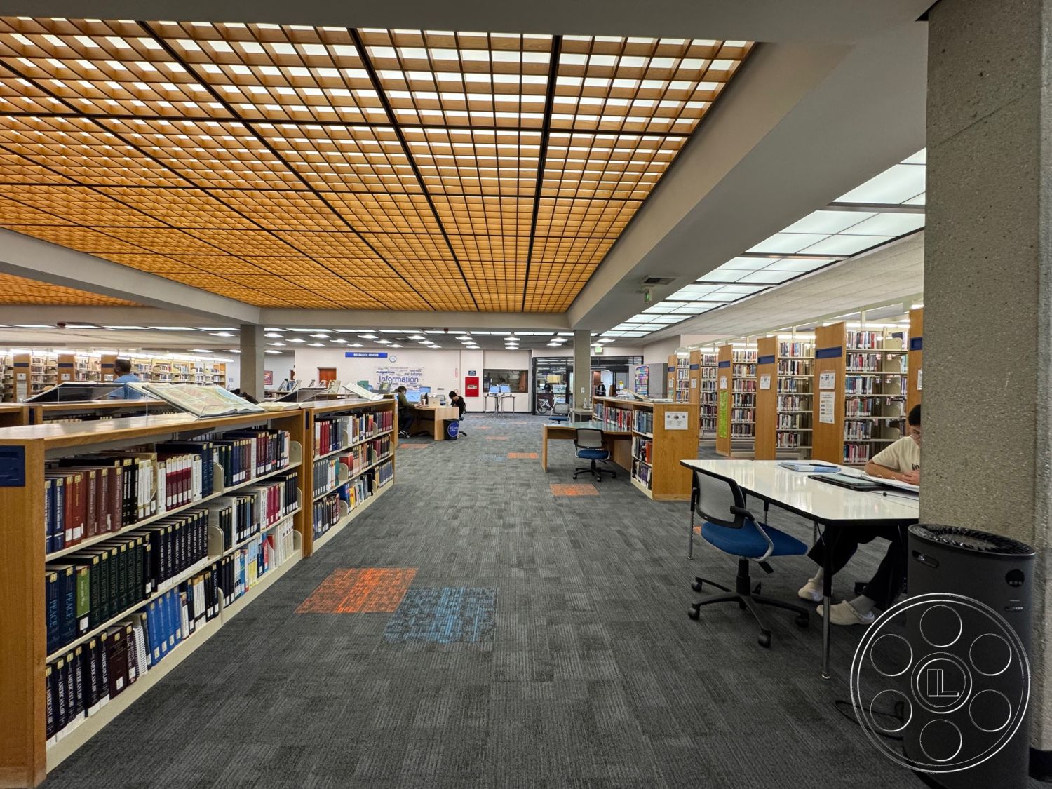 School 3 - bright natural lighting, acoustic ceiling tiles, textured carpet flooring, flush-mounted LED lighting, interior library space, wooden library shelving units, exposed concrete support columns, open reading areas, collaborative study tables, library information desk, modern ergonomic seating, grid-patterned wood ceiling, designated quiet zones, colorful carpet inlay designs