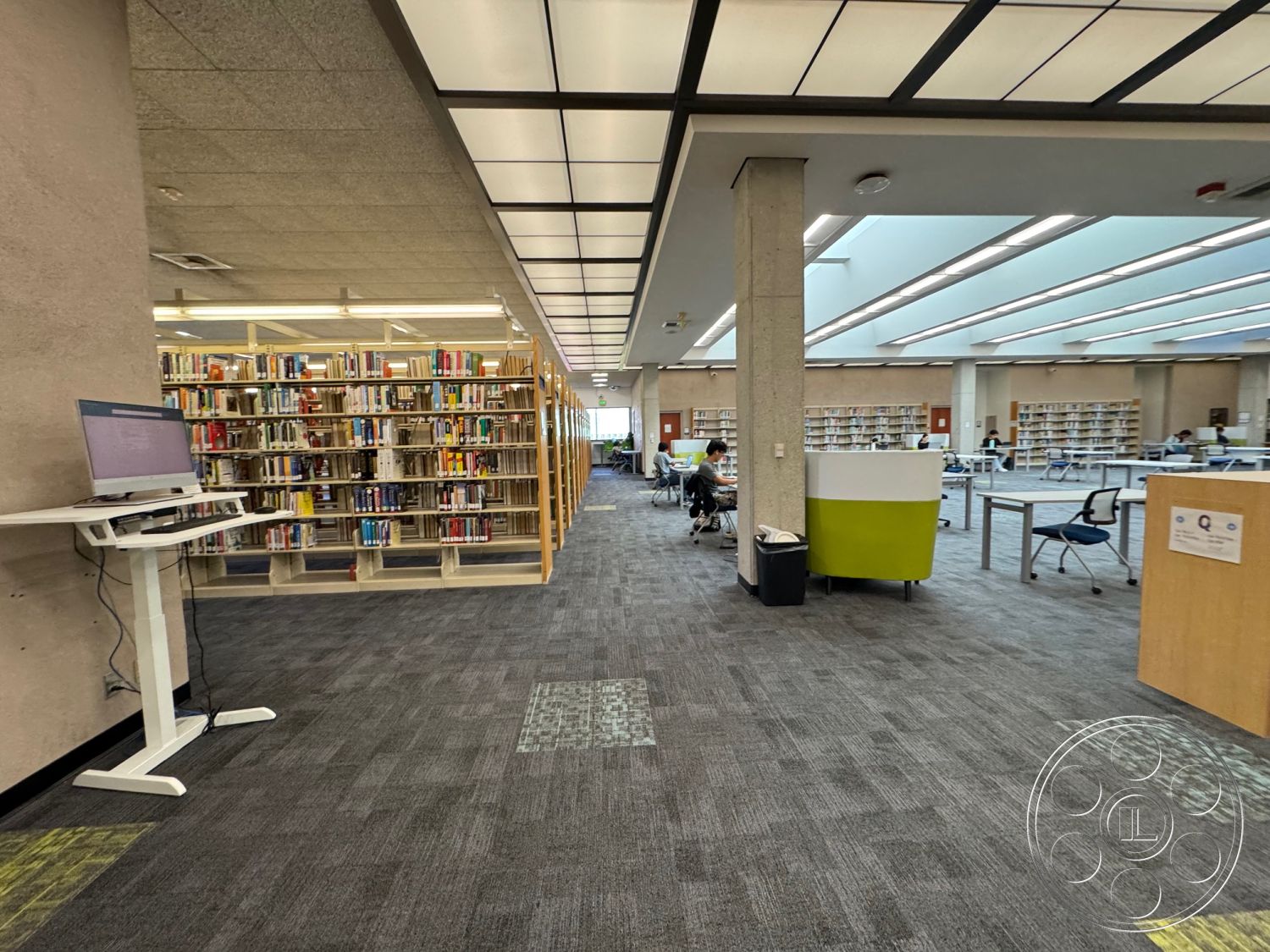 School 3 - neutral color palette, open floor plan, interior space, concrete columns, ergonomic chairs, overhead fluorescent lighting, natural light from skylights, collaborative workspaces, floor carpet tiles, modular study desks, wooden bookcases, angled ceiling design, glass windows for visibility, public library setting, quiet reading areas, community-accessible interior, modern computer workstation