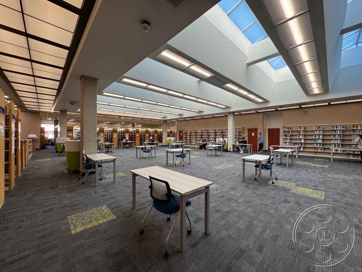 School 3 - carpet flooring, textured wall paneling, community gathering space, accessible layout, LED overhead lighting, concrete support columns, interior library space, flexible seating arrangements, open ceiling design, large skylights, light wood shelving units, minimalist study tables, adjustable swivel chairs, spacious reading area, modern library aesthetic