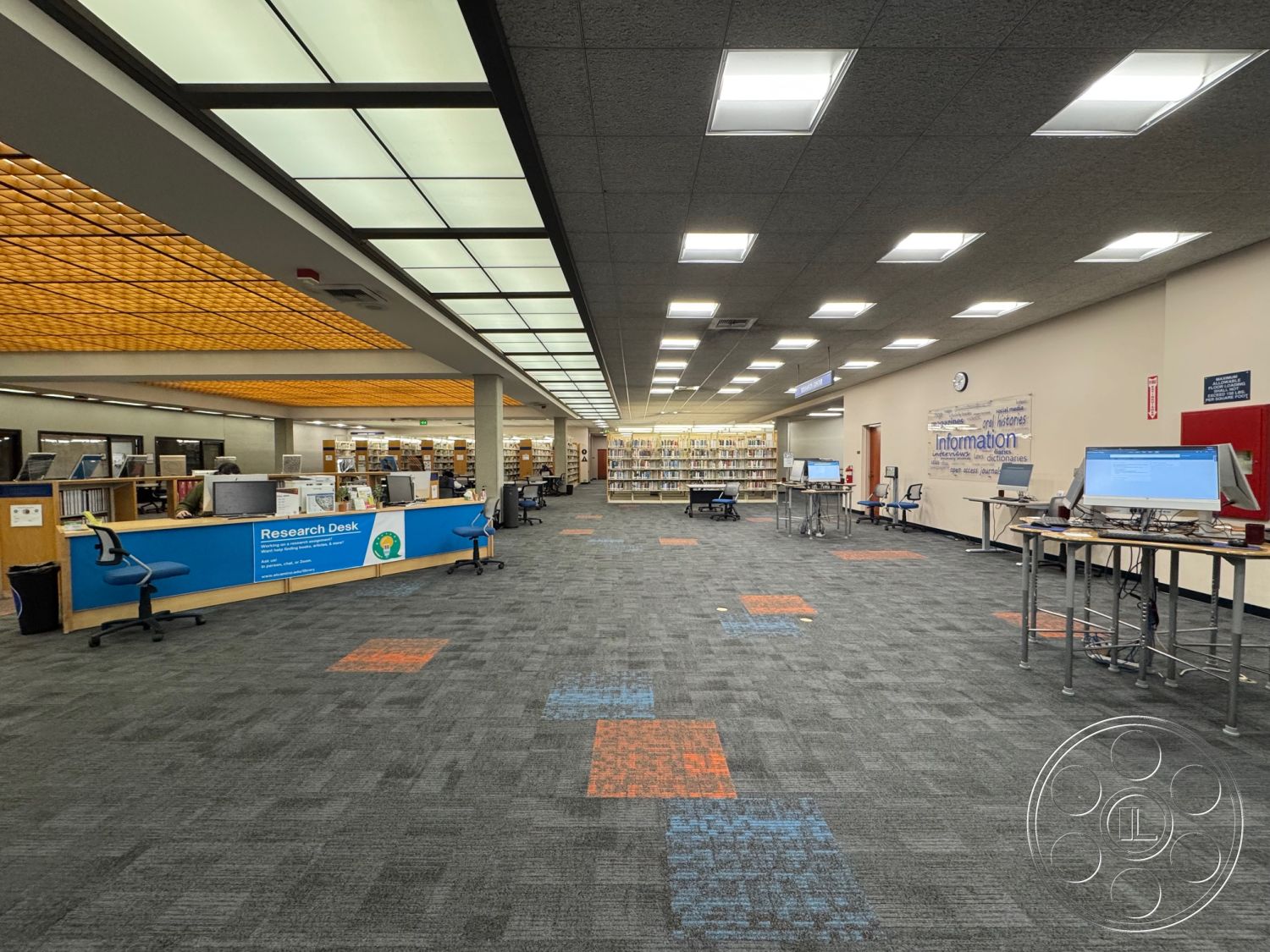 School 3 - modern lighting fixtures, textured wall finishes, contemporary furniture design, neutral painted walls, interior library space, shared workspace environment, carpet flooring with colorful tile accents, drop ceiling with integrated lighting panels, open study areas with computer stations, blue laminate research desk, wooden book shelving units, task chairs in blue upholstery, information signage wall, high ceiling with acoustic tiles, library circulation desk area