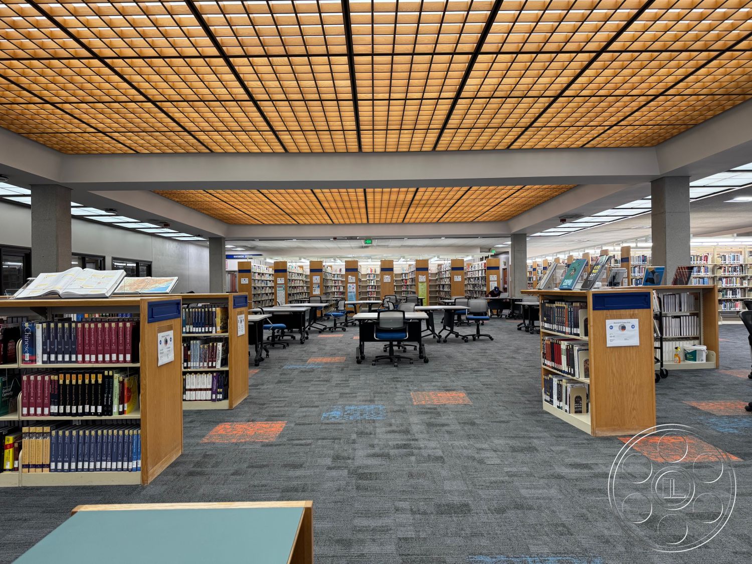 School 3 - acoustic ceiling tiles, open floor plan layout, textured carpet flooring, linear LED ceiling lights, modern office chairs, metal trash receptacles, coffered ceiling design, wooden bookcase shelving units, interior library space, exposed concrete columns interior, library study tables, blue and orange carpet tiles, glass windows for natural light, light grey painted walls, flexible seating areas, commercial grade carpeting, public reading area, informational signage boards