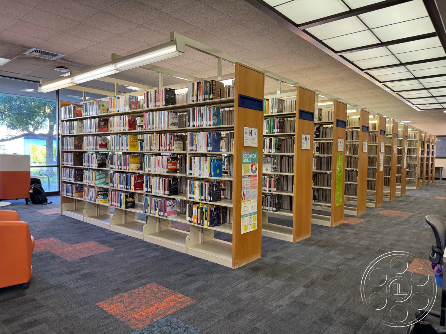 School 3 - overhead fluorescent lighting, collaborative workstations, interior library space, carpet flooring with patterned accents, open shelving units with wooden frames, modular seating in orange and white, glass windows with views to exterior landscape, study area with ergonomic office chairs, promotional posters on shelving units, wooden bookends for organization, natural daylight from large windows, reading nook with comfortable seating