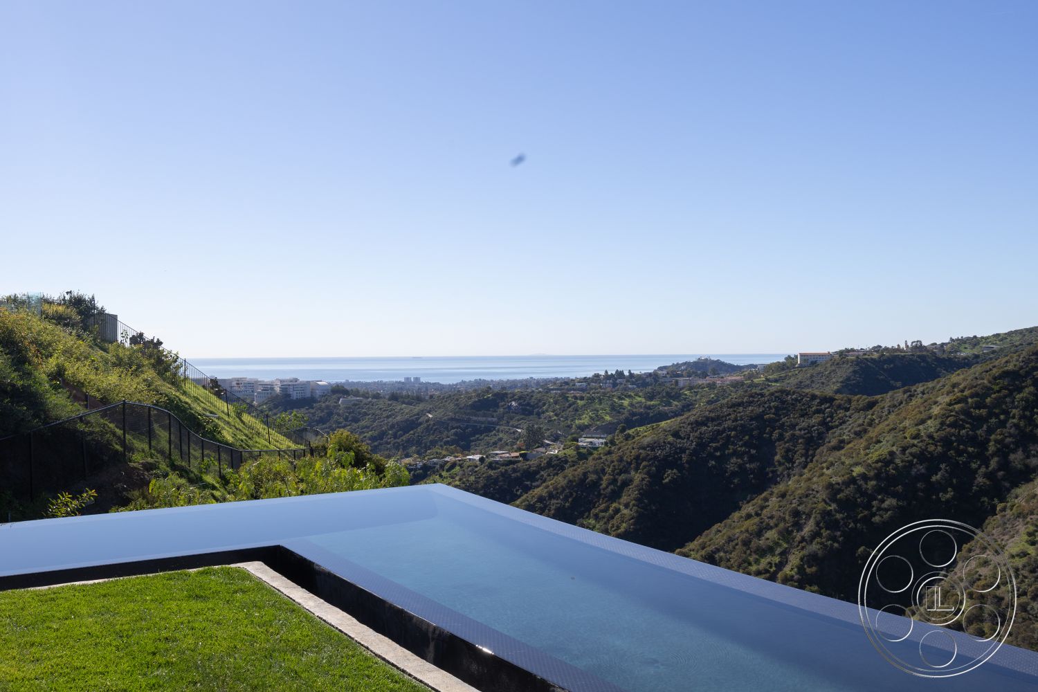 Modern 516 - infinity pool with black tile edge, sloped hillside landscape with lush greenery, concrete pool deck with grass border, ocean view from hillside property, fenced backyard with black metal railing, modern architecture with expansive vistas, outdoor living space with scenic backdrop, low-maintenance landscaping among rocky terrain, rectangular infinity edge pool, exterior space with natural elements