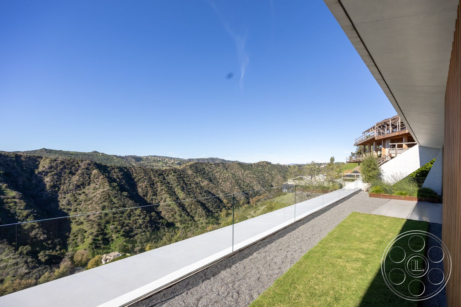 Modern 516 - natural stone accents, open outdoor space, green grass lawn, minimalist landscaping, hillside location, smooth concrete patio, flat roof design, wooden deck area, modern architectural design, exterior glass balcony railing, concrete outdoor walkway, gravel landscape garden, large glass windows with panoramic views, blending indoor and outdoor living, grazing land view, contemporary hillside residence, mountain range backdrop