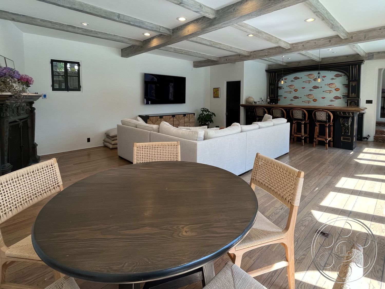 Medit 166 - natural light, wooden bar stools, cozy atmosphere, light wood flooring, open-concept living space, hardwood floor, flat-screen television, plant decoration, woven dining chairs, round wooden dining table, exposed wood ceiling beams, white painted walls, modern sectional sofa, black fireplace surround, bar area with fish mural