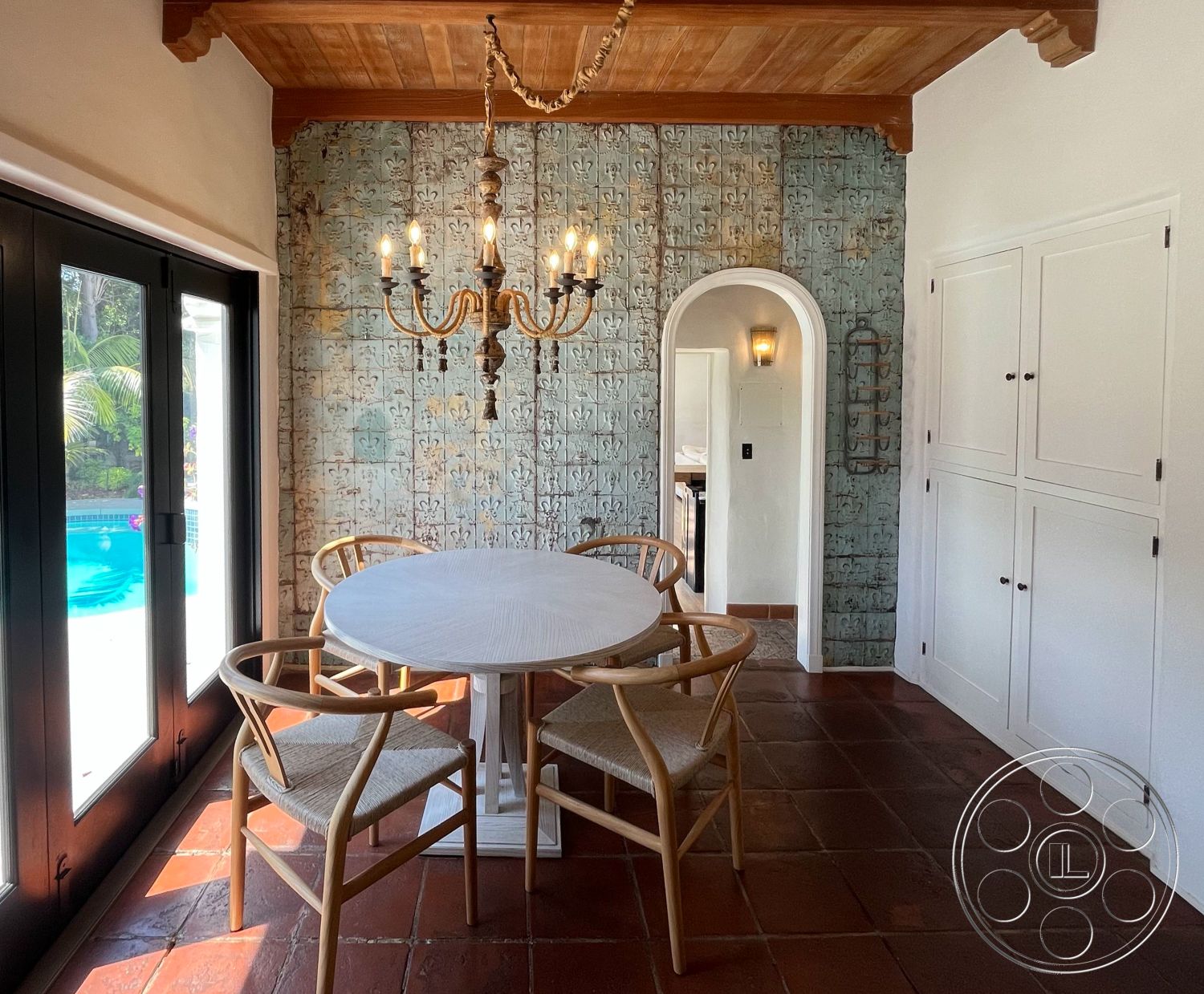 Medit 166 - arched doorways, textured plaster walls, poolside view, earth-toned color palette, interior dining area, natural light-filled space, built-in white cabinets, exposed wood ceiling, chandelier lighting fixture, clay tile flooring, white wooden dining table, woven seating chairs, decorative tiles with floral pattern, large glass doors to pool area, rustic Mediterranean style