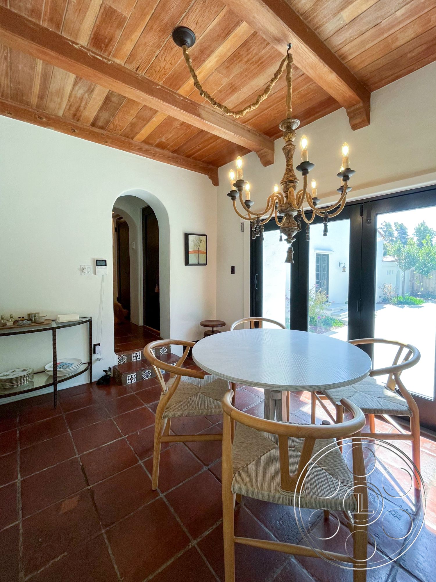 Medit 166 - open concept design, exposed wood beams, casual dining area, round dining table, iron chandelier, arched doorways, white plaster walls, woven dining chairs, vaulted wood ceiling, rustic Mediterranean style, terrazzo tile flooring, wood cabinetry accent, decorative ceramic tile steps, natural light from sliding doors, dining nook with garden view, sunlit breakfast area, home with indoor-outdoor flow