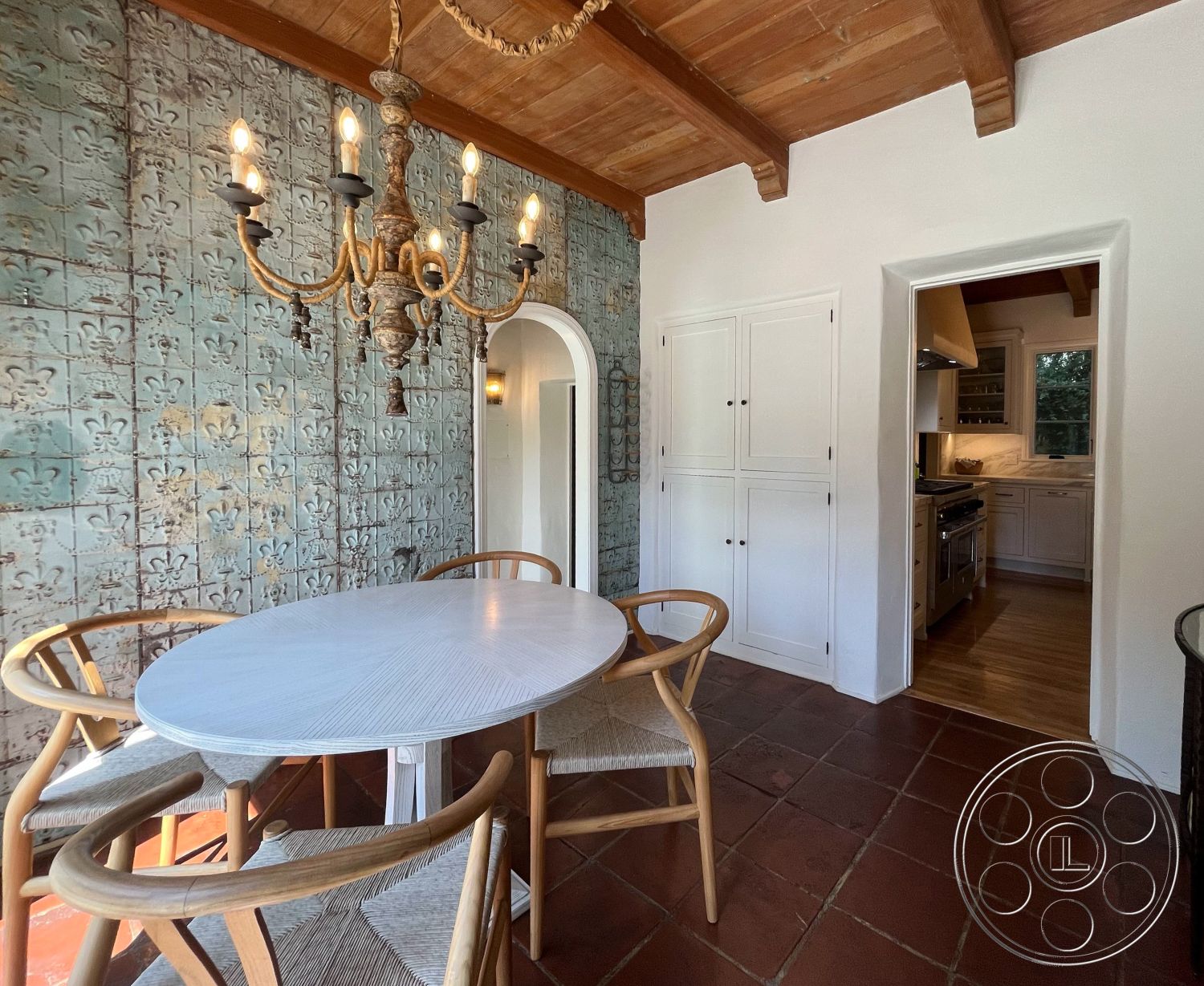 Medit 166 - natural wood cabinetry, earthy color palette, arched doorway, Mediterranean architectural style, terra cotta floor tiles, exposed wood ceiling beams, oval wooden dining table, open floor plan dining area, ceramic tile wall treatment, woven chair seats, white painted kitchen walls, wall sconce lighting, antique chandelier, rustic interior, kitchen access via archway, interior decorative elements