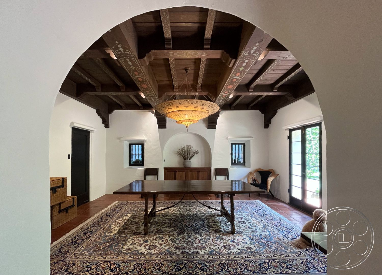 Medit 166 - wood dining table, ceramic tile flooring, textured plaster walls, decorative wall accents, exposed wood ceiling beams, vintage area rug, dark wood window frames, natural light through glass doors, traditional chandelier lighting, Spanish-style arched doorways, black door hardware, woven baskets storage