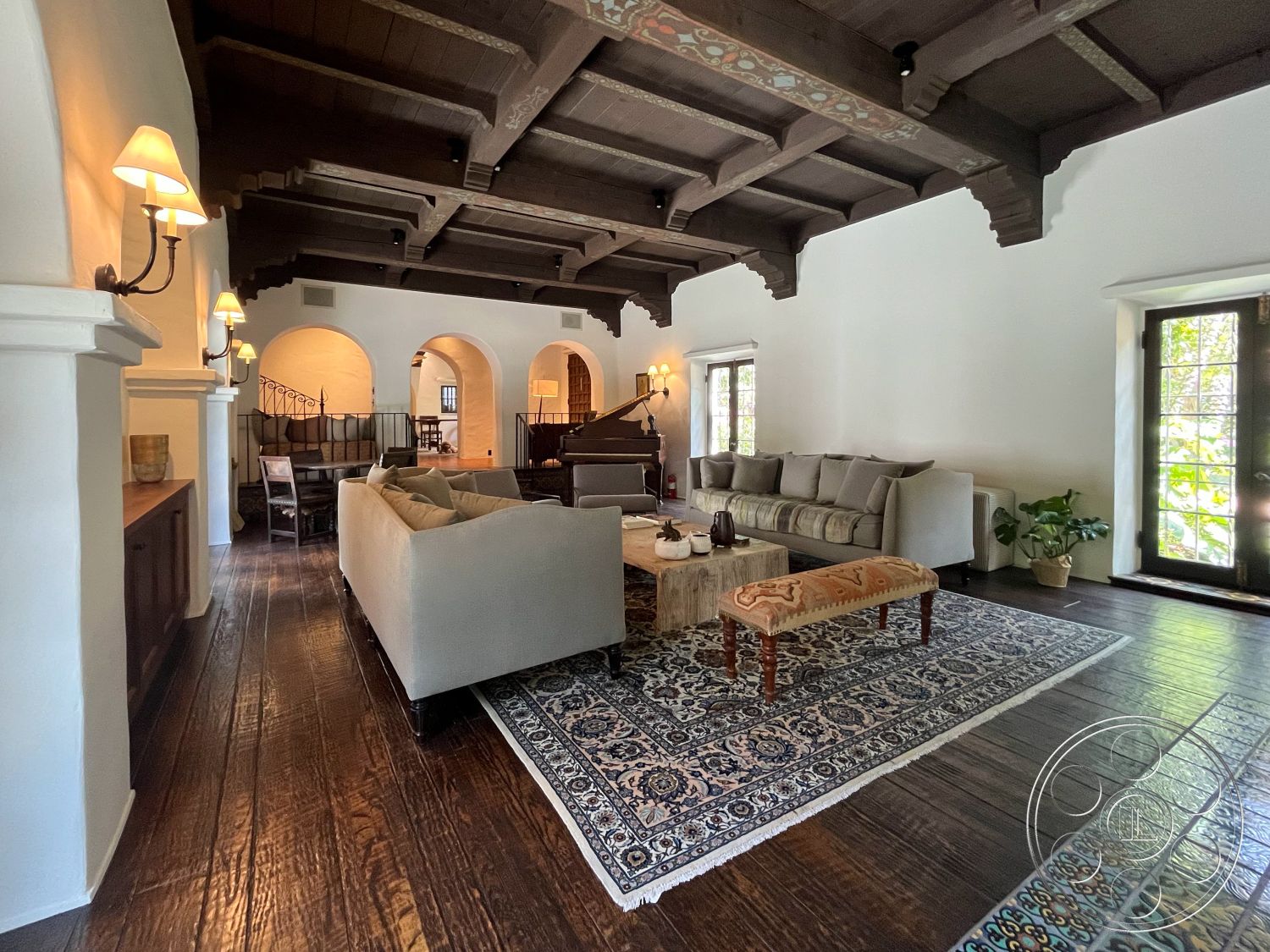 Medit 166 - plant decor, wooden coffee table, open floor plan, brick fireplace, arched doorways, textured plaster walls, Mediterranean architectural style, exposed wood ceiling beams, hardwood living room floors, traditional area rug, white painted walls, upholstered sectional sofa, dark wood trim details, vintage accent bench, dual-pane windows, interior lighting sconces, decorative ceiling molding, shaded outdoor patio view