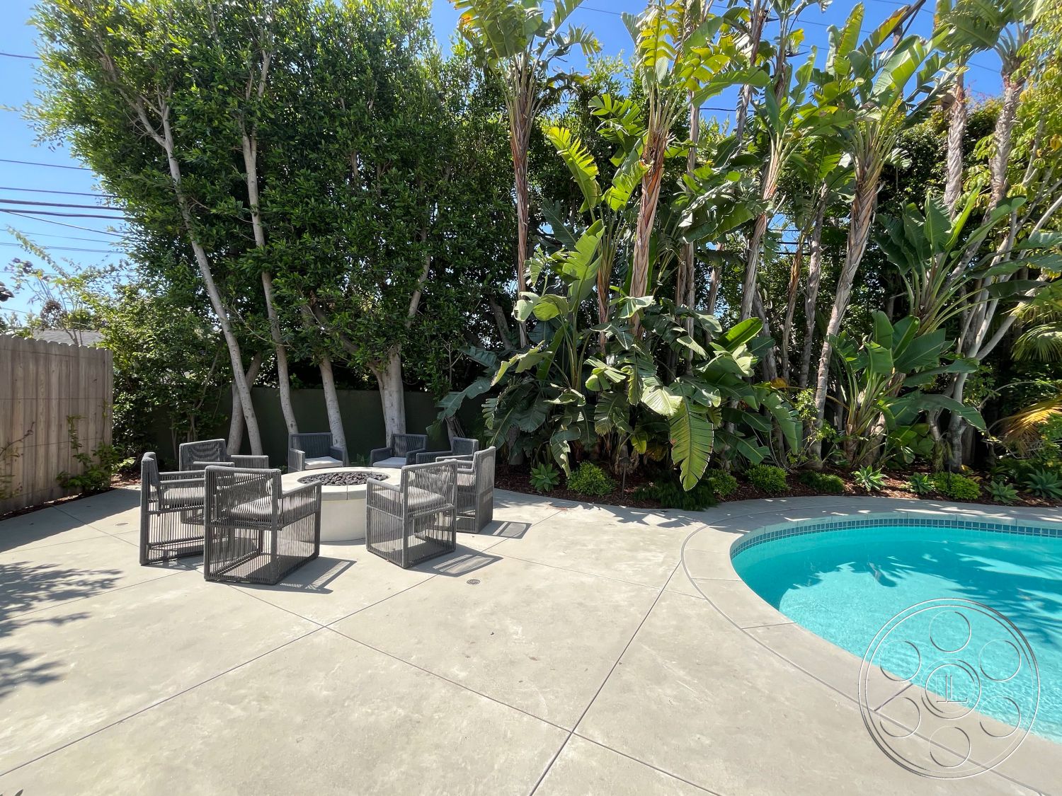 Medit 166 - lush tropical landscaping, wooden privacy fence, surrounding palm trees, metal outdoor chairs, oval swimming pool, sunny backyard setting, exterior concrete patio flooring, concrete pool edge, modern outdoor living space, fire pit seating area