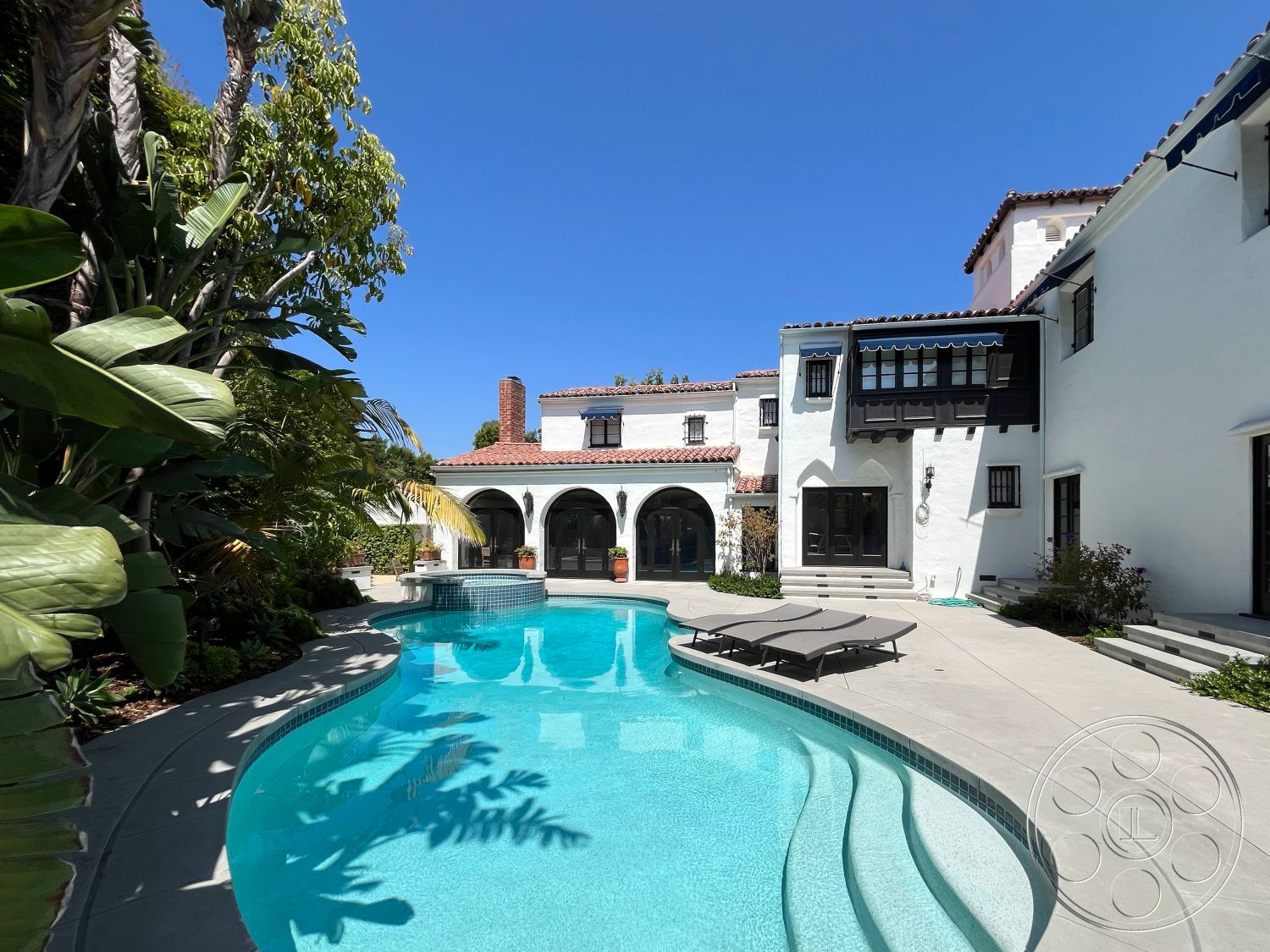 Medit 166 - exterior, paver driveway, exposed wood beams, outdoor living space, tropical landscaping, arched doorways, tile roof, Mediterranean architectural style, blue water pool, curved pool shape, landscaped garden area, stucco exterior walls, concrete patio floor, stone planter beds, built-in outdoor seating, ceramic tile pool border, black double-hung windows, sun loungers beside pool, poolside oasis