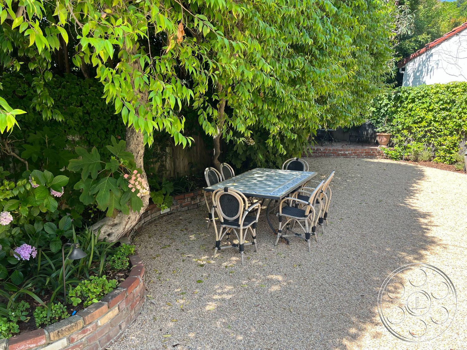 Medit 166 - textured stone wall, surrounding greenery, outdoor dining area, glass-top dining table, lush green landscaping, metal dining chairs, exterior garden patio, garden lighting fixtures, private backyard setting, brick seating wall, gravel patio floor, overhanging tree canopy, flower bed with hydrangeas, curved patio layout
