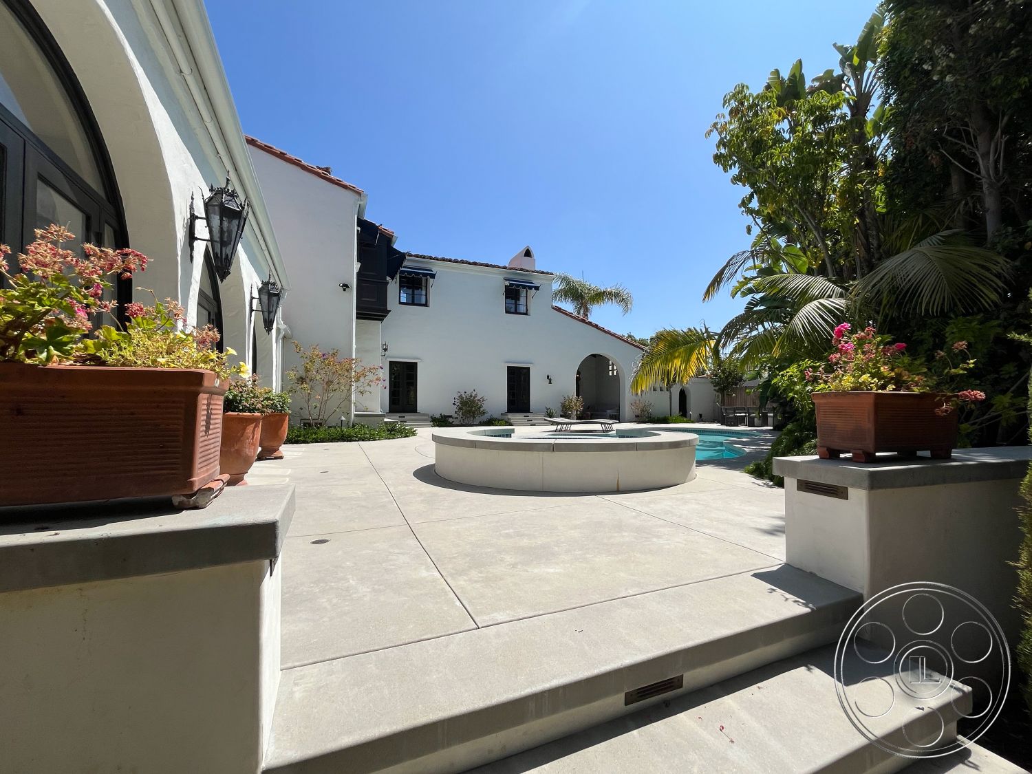 Medit 166 - white stucco walls, potted plants, outdoor seating area, modern landscaping, clear blue sky, wooden pergola, arched doorways, suburban backyard, rectangular pool, stone steps, Mediterranean architectural style, concrete patio floor, tile pool deck, exterior lantern sconces, multicolored flower pots