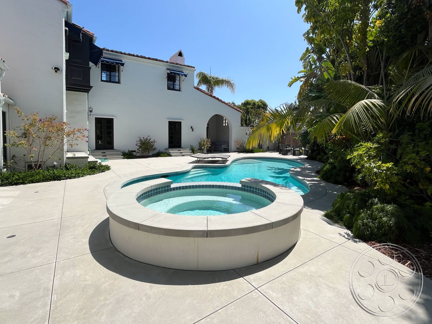 Medit 166 - exterior, rectangular swimming pool, arched entryway, lush tropical landscaping, concrete pool deck, outdoor relaxation area, curved pool shape, modern outdoor seating area, sunny outdoor environment, white stucco exterior walls, round spa with tile accents, textured concrete patio surface, palm tree surrounded setting, urban backyard oasis, California-style home