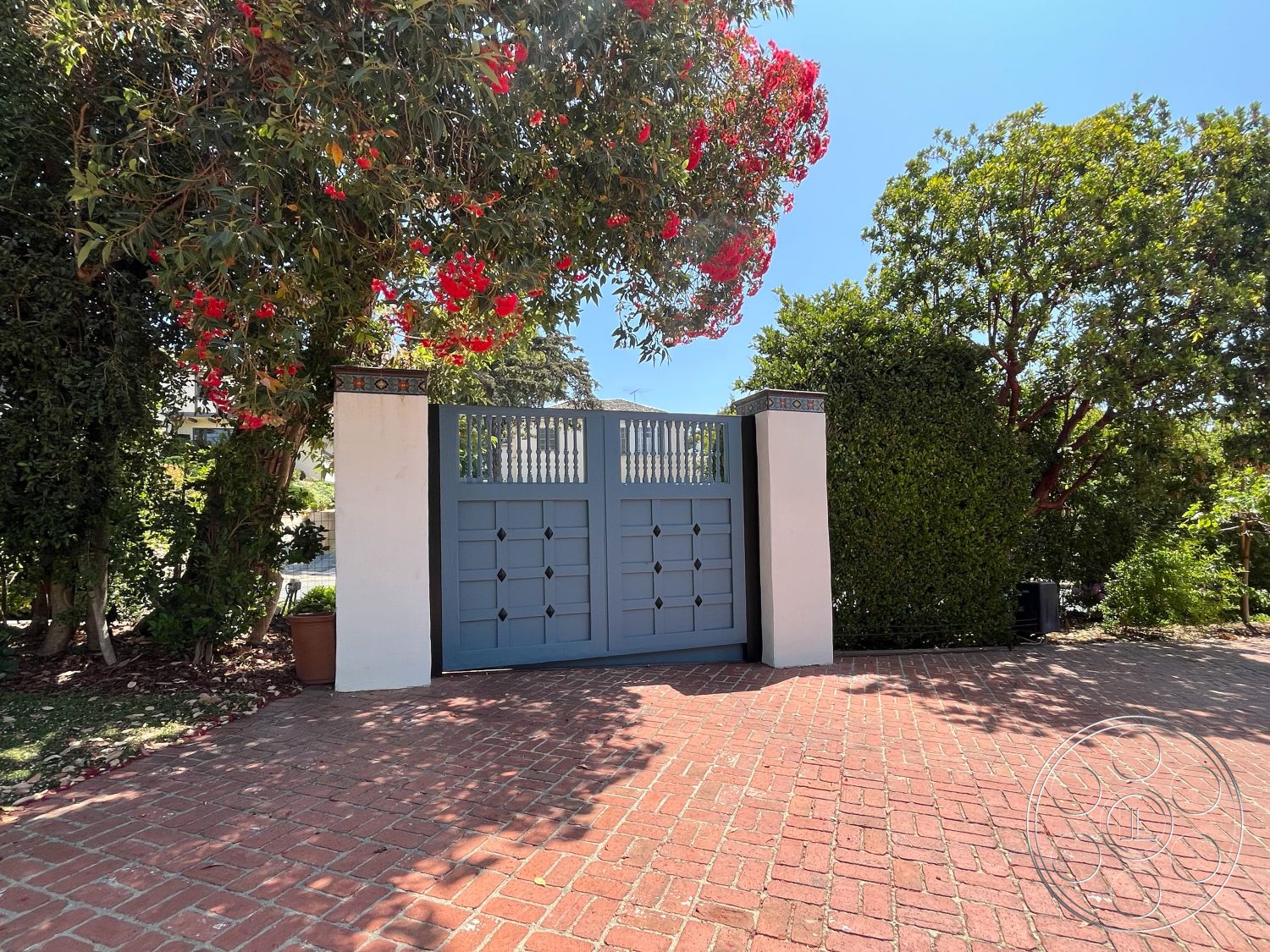 Medit 166 - landscaped garden setting, urban residential environment, exterior gate design, blue wooden double gates, bricked driveway entrance, lush leafy greenery, flowering tree canopies, ornamental brick pattern, textured white stucco walls, tiled decorative accents, outdoor privacy hedges, bright sunlight casting shadows