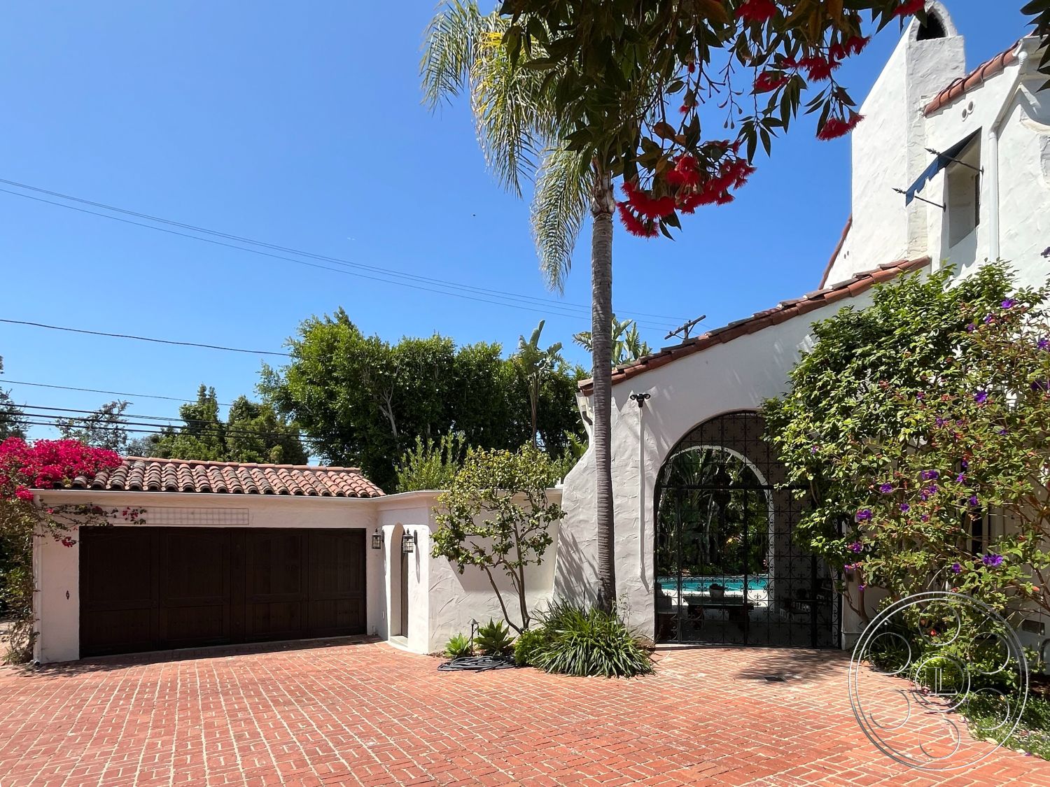 Medit 166 - exterior, palm trees, two-car garage, lush tropical landscaping, Spanish-style architecture, corner lot, urban residential setting, clay tile roof, courtyard access, archway entrance, stucco exterior walls, brick driveway pavers, decorative wrought iron gates, bougainvillea plants, quaint garden area, outdoor pool area visible, sunny blue sky, wooden garage doors