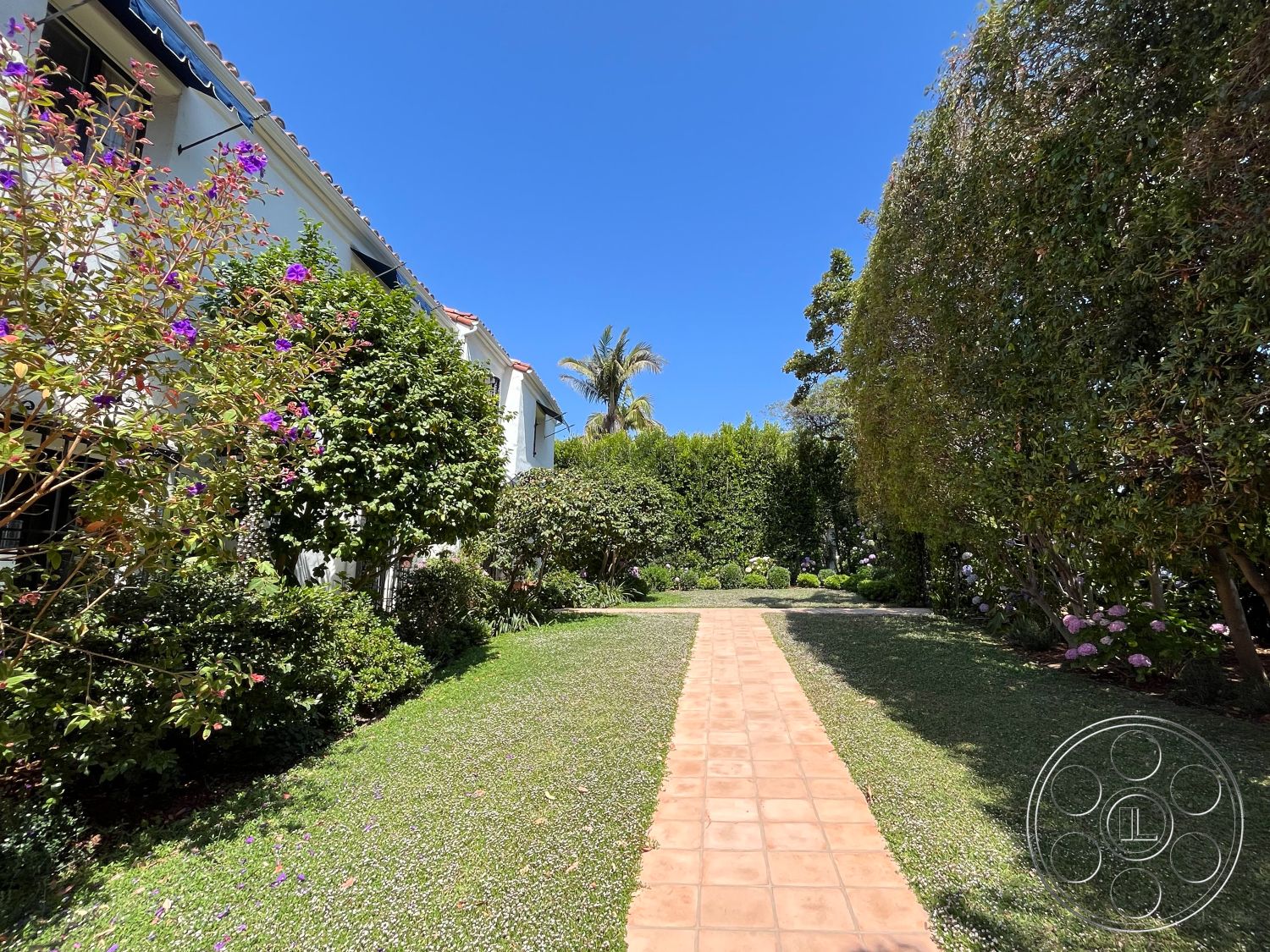 Medit 166 - white stucco walls, flowering shrubs, green hedges, tropical palm trees, exterior garden pathway, manicured lawn area, terracotta tile walkway, lush landscaping with flowering plants, city/suburban residential setting, sloped roof with terracotta tiles, outdoor greenery around entrance, bright blue sky background, residential garden design, symmetrical pathway layout, drought-tolerant plantings