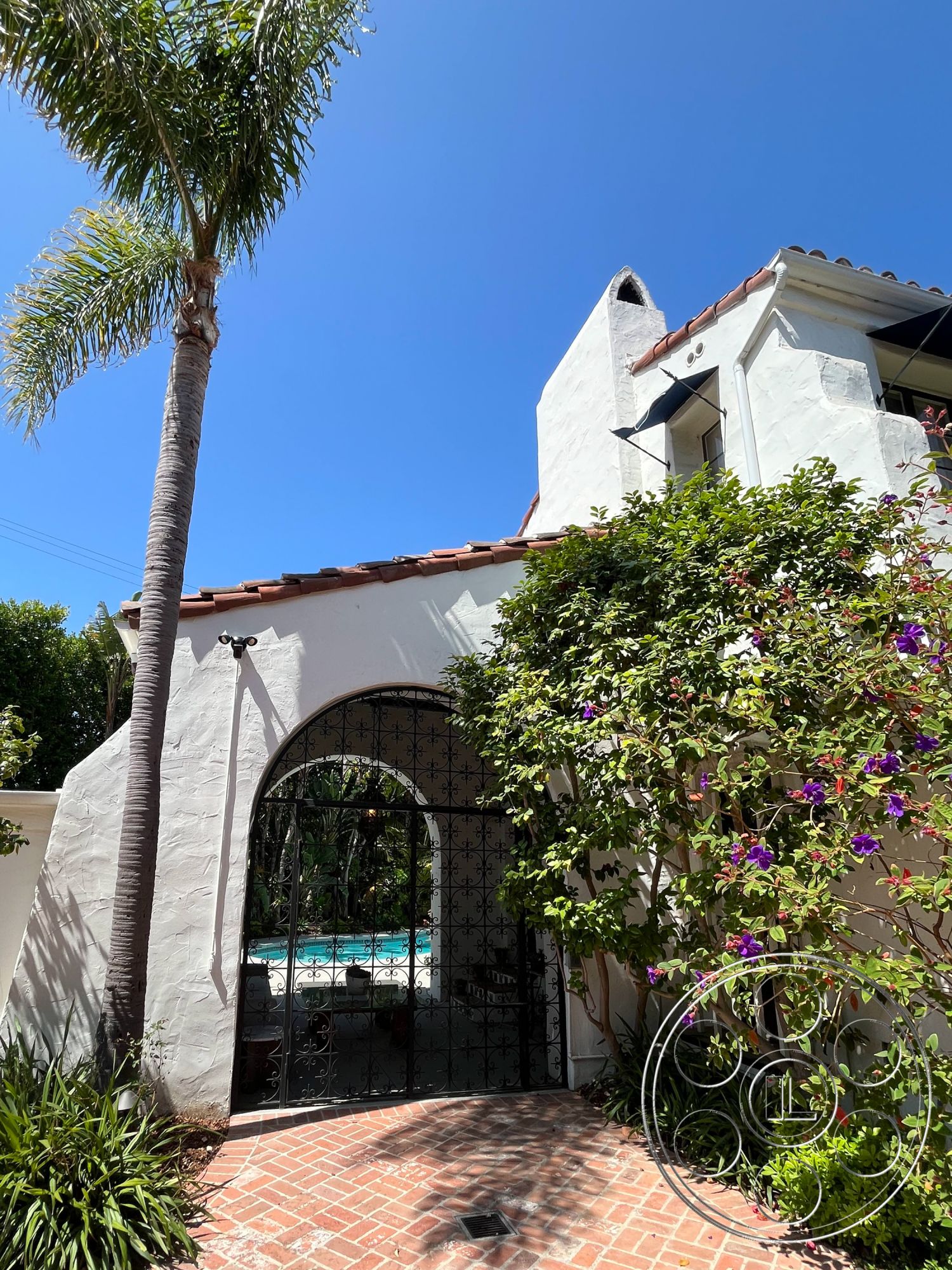 Medit 166 - exterior, palm trees, tropical landscaping, arched doorways, textured plaster walls, red clay tile roof, Mediterranean architectural style, sunny outdoor setting, stucco exterior walls, lush green shrubbery, arched wrought iron gate, bricked pathway, flowering vines, pool view from gate