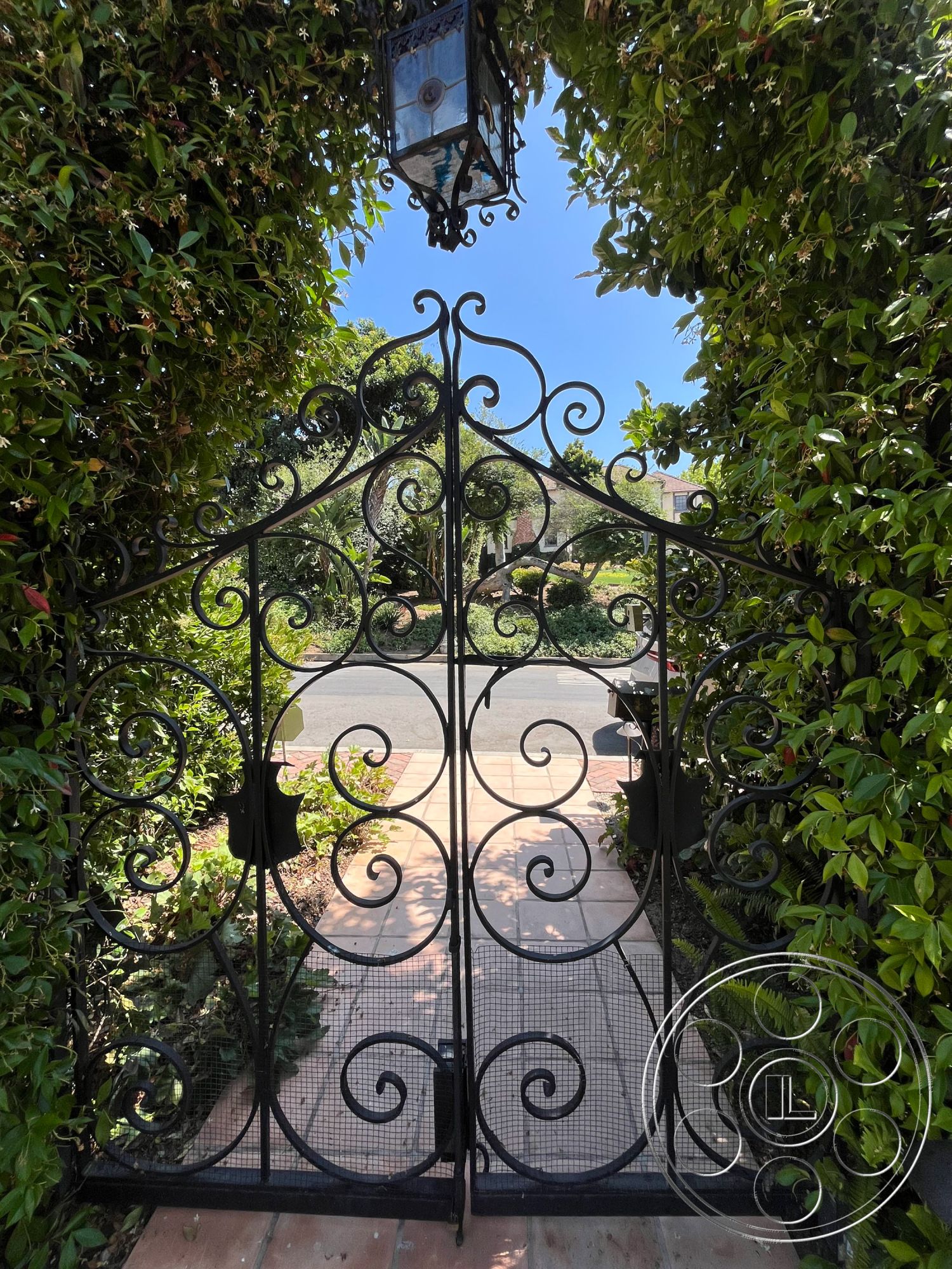 Medit 166 - urban garden setting, wrought iron gate with ornate design, exterior entrance gate, landscaped garden entrance, paved walkway with terracotta tiles, arched gate design, framed by lush green foliage, traditional lantern fixture, arched threshold with view of exterior pathway, textured foliage surrounding entrance, welcoming garden pathway, decorative ironwork details, soft sunlight filtering through greenery