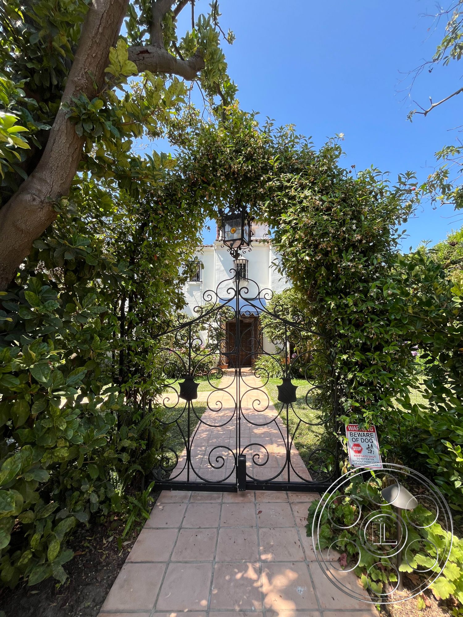 Medit 166 - landscaped garden, white stucco walls, traditional architecture, urban garden, flowering plants, residential setting, suburban backyard, arched entrance, hedge borders, sunlit exterior, exterior lush greenery, hardscaped walkway, wrought iron garden gate, tile pathway, wooden door with decorative arch, brick accent in pathway, courtyard style, ornate gate design