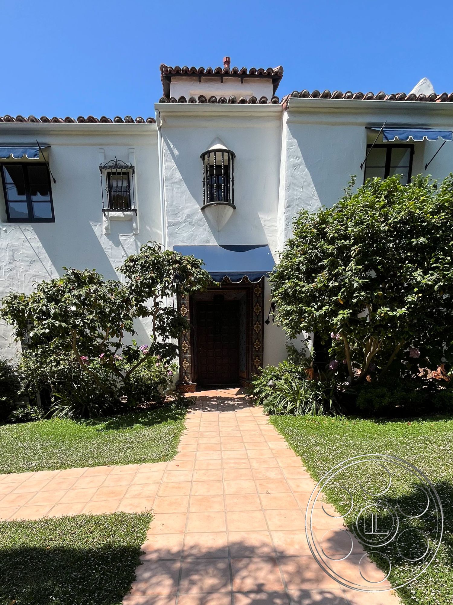 Medit 166 - exterior, landscaped garden, lush greenery, curved pathway, planted shrubs, sunny setting, stucco walls, arched doorways, decorative tile accents, terra cotta roof tiles, wooden front door, Mediterranean architectural style, ornate metal light fixture, terracotta walkway